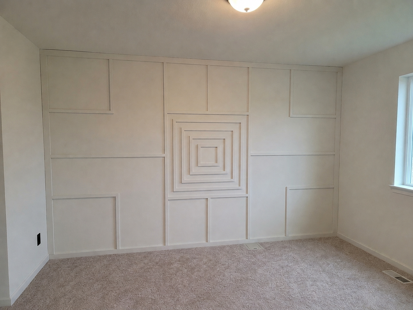 Empty room with beige carpet and white walls, featuring a geometric recessed panel wall design in the center.