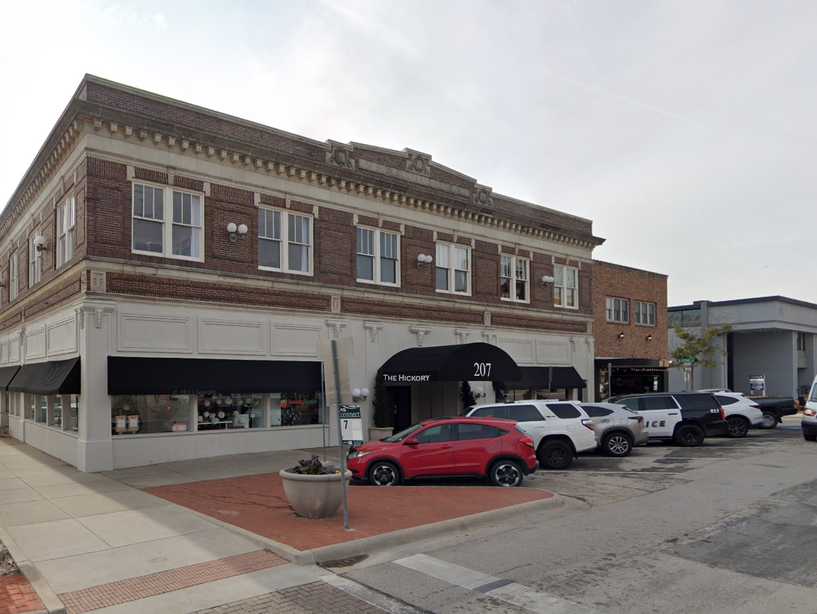 Street view of a corner brick building with shops on the ground floor, black awnings, and white upper floors with multiple windows. Several cars are parked in front, including a red car and police cars. There is a small sidewalk with a planter and a parking sign.