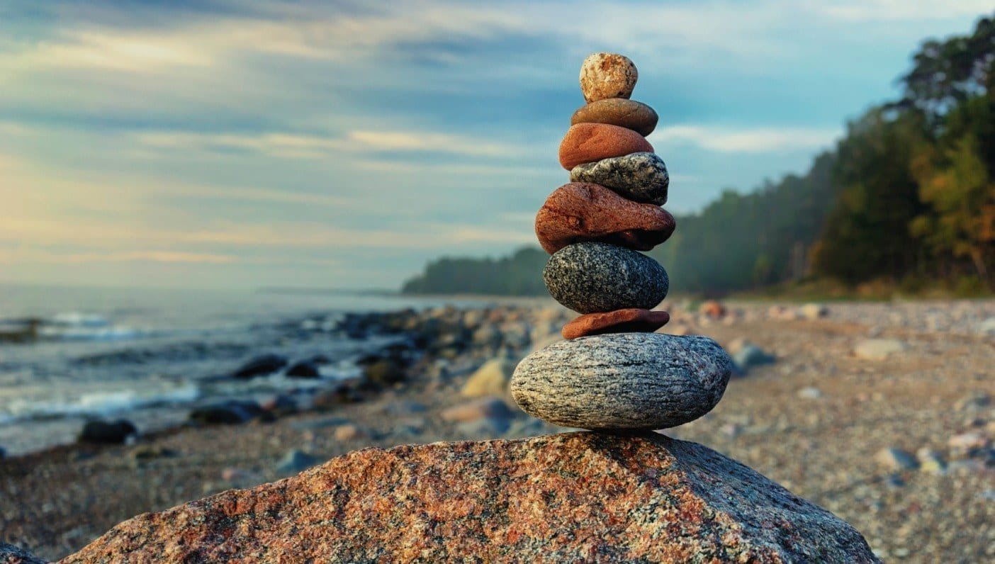 Stacked rocks on a rock at the beach with the ocean and trees in the background, under a partly cloudy sky.