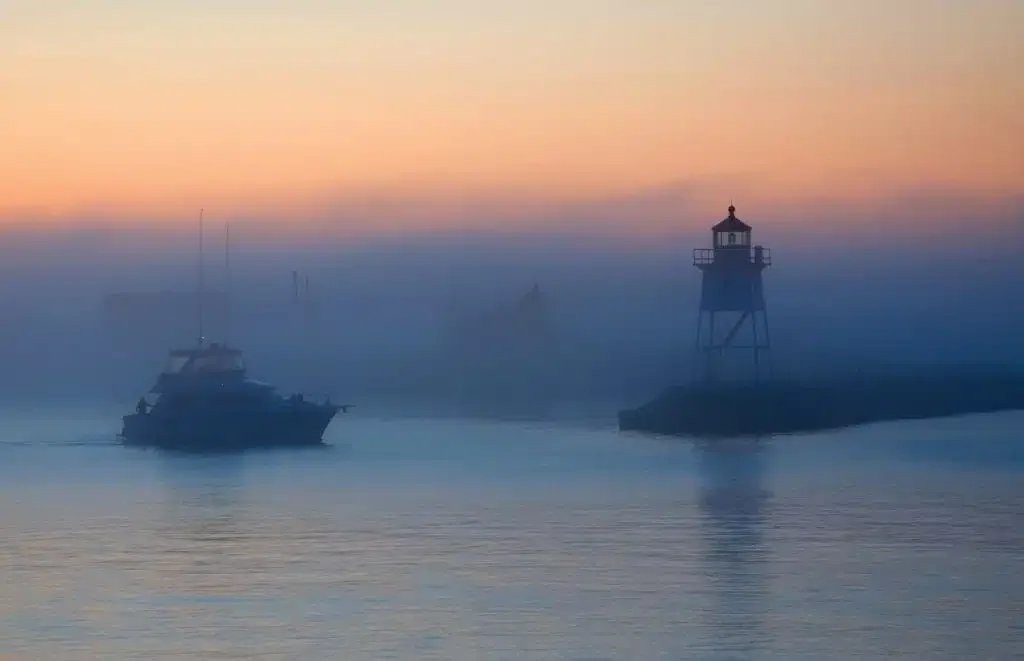 A boat sailing near a lighthouse during foggy sunrise or sunset.