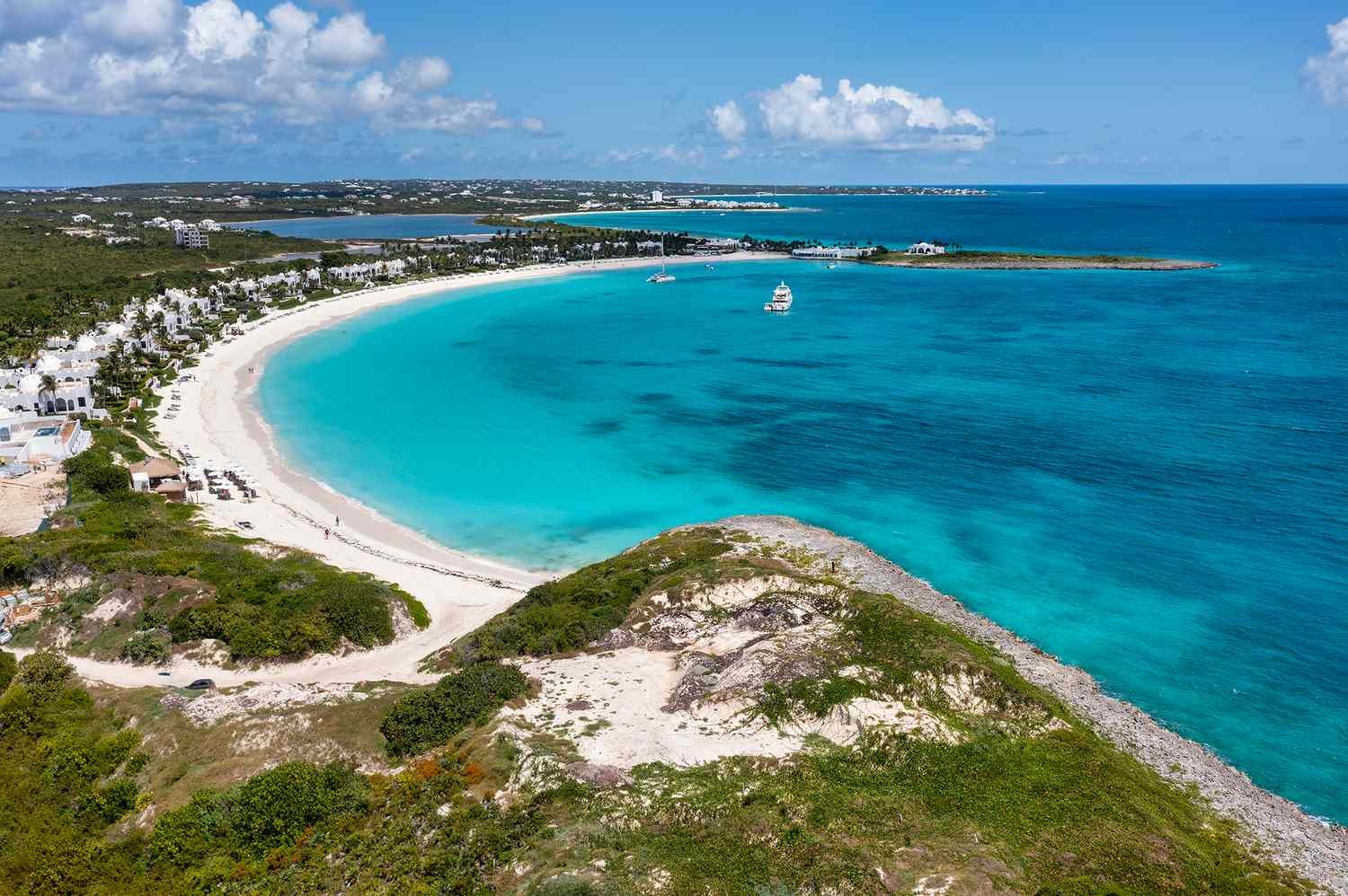 Aerial view of a scenic Caribbean beach with white sandy shores, turquoise waters, lush greenery, and boats anchored near the shore.