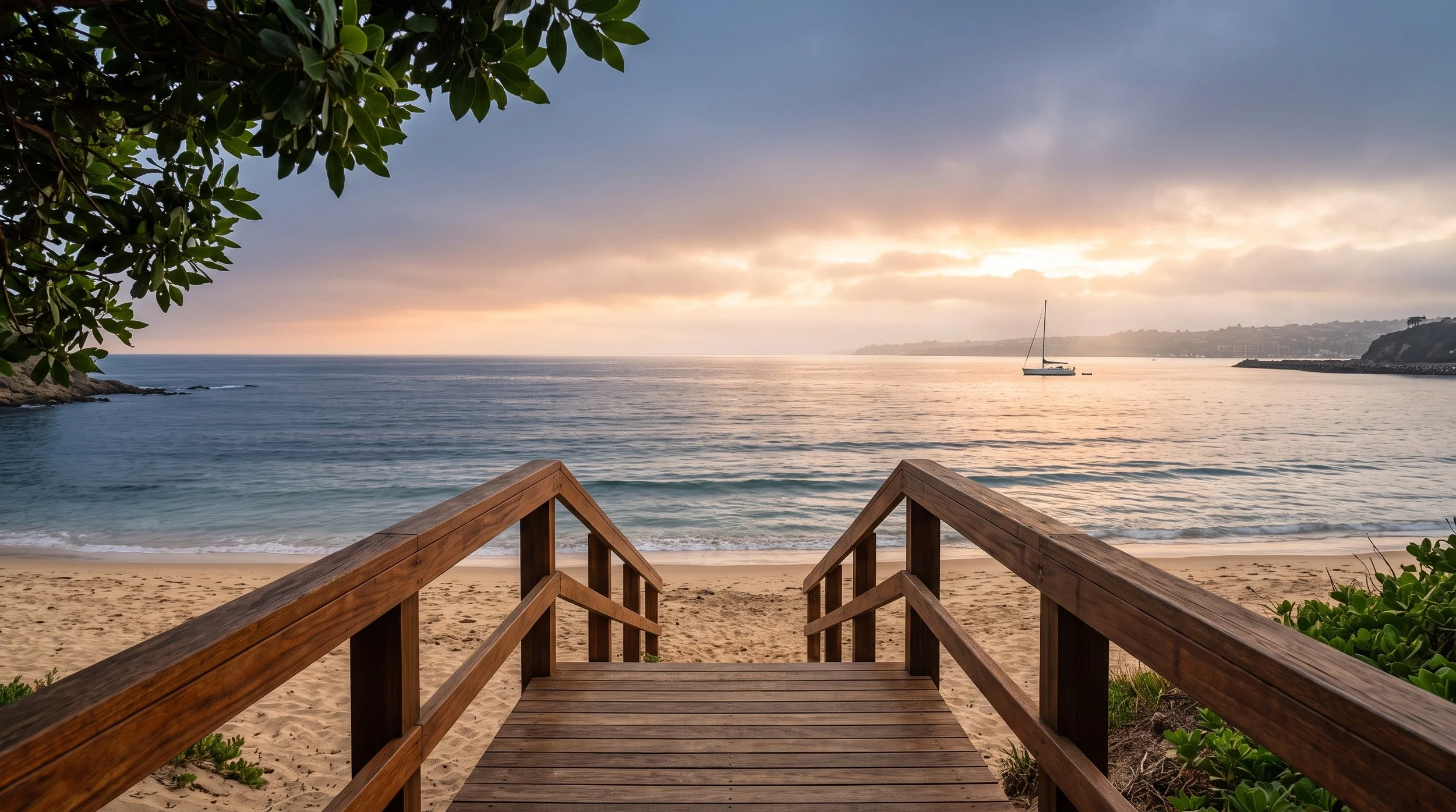 Wooden staircase leading down to a sandy beach, overlooking calm ocean waters at sunset, with a boat anchored offshore and a cloudy sky.