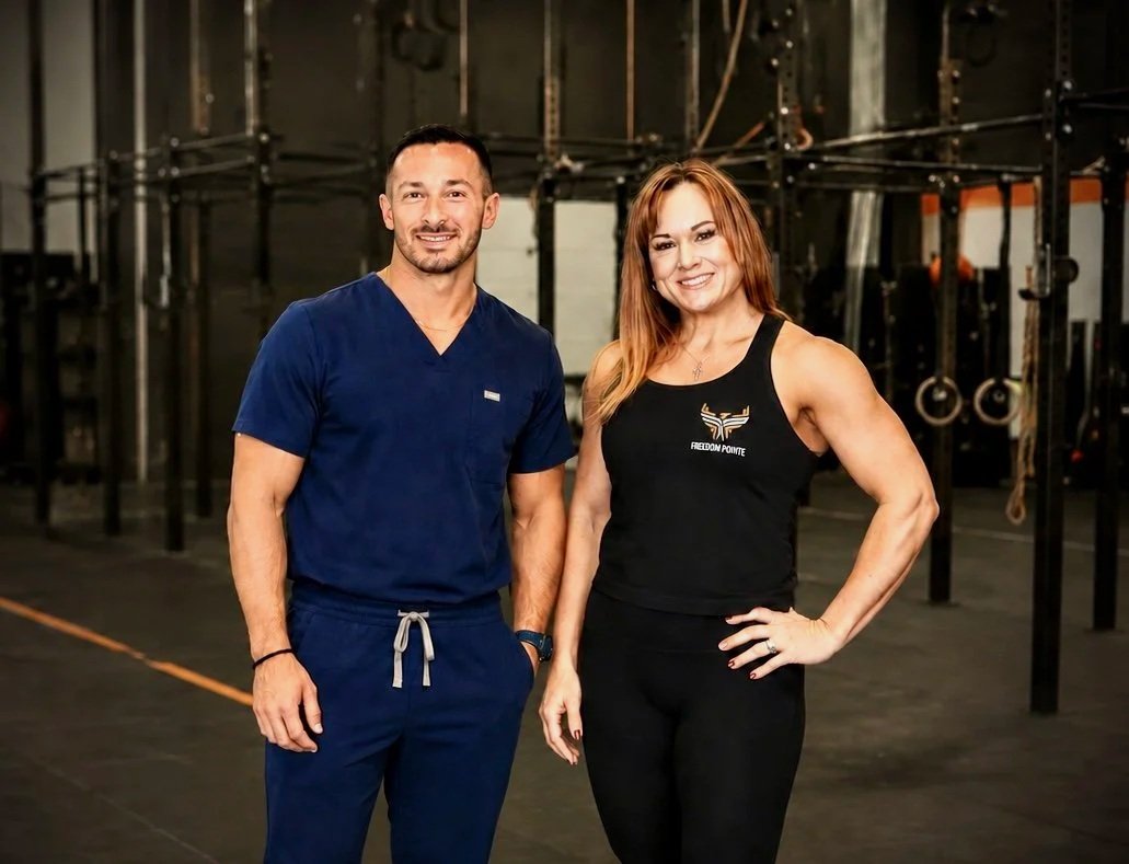 A man in blue medical scrubs and a woman in black athleticwear standing in a gym with fitness equipment in the background.