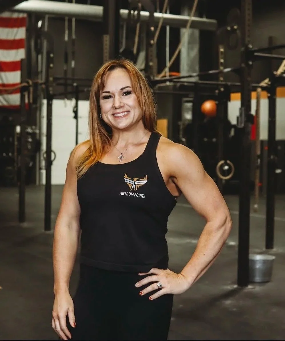 A woman with red hair and a muscular build standing in a gym, wearing a black tank top with a logo that says 'FREEDOM POINTE'. She is smiling, with her right hand on her hip. The gym background includes equipment like ropes, rings, and an American flag.