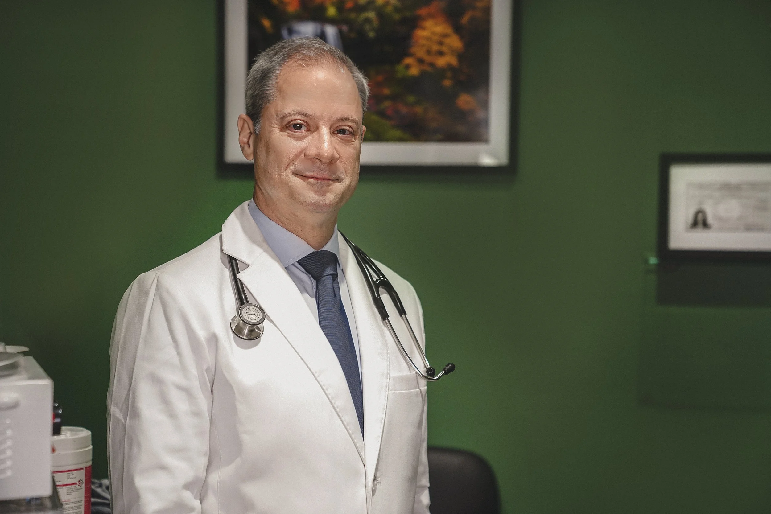 Doctor William Berger, wearing a white lab coat, a stethoscope around his neck, and a blue tie, standing in a medical office with green walls and framed pictures in the background in Northeast Medical Practice.