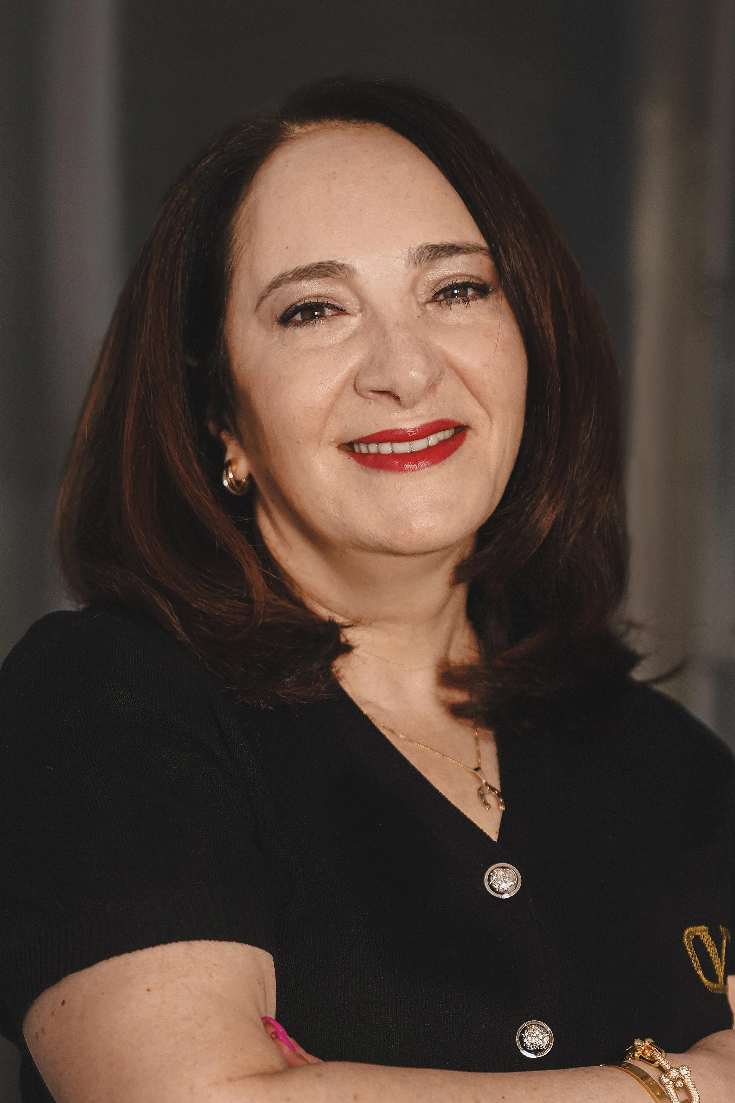 Irina Stainov with shoulder-length brown hair smiling, wearing red lipstick, earrings, a black top with buttons, and jewelry including a bracelet and necklace, with arms crossed.