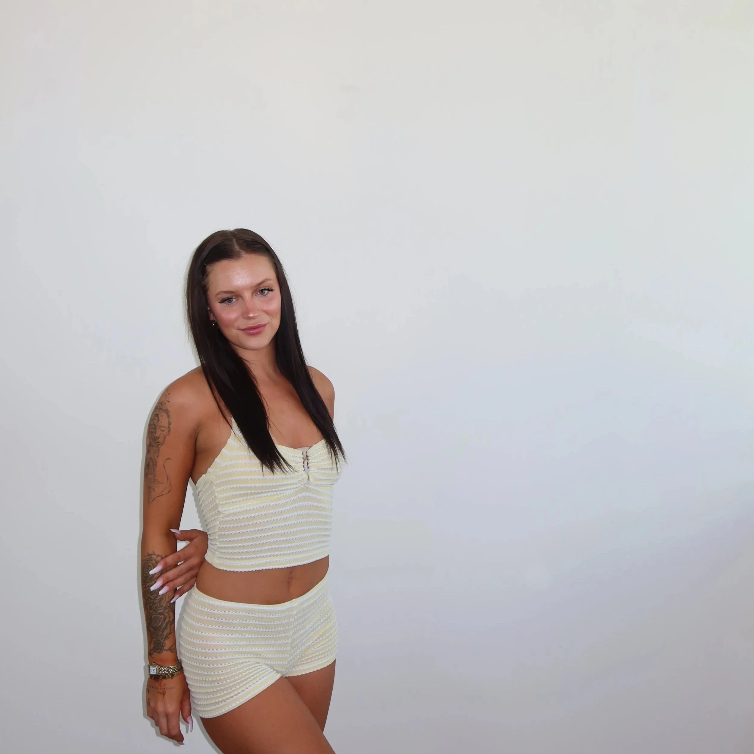A woman with long dark hair wearing a yellow and white striped top and matching shorts standing against a plain white background.
