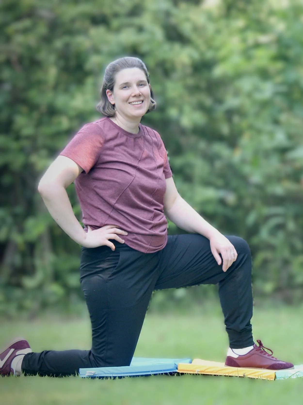 Woman doing a lunge exercise outdoors on grass with a green blurred background, wearing a maroon shirt, black pants, and maroon sneakers.