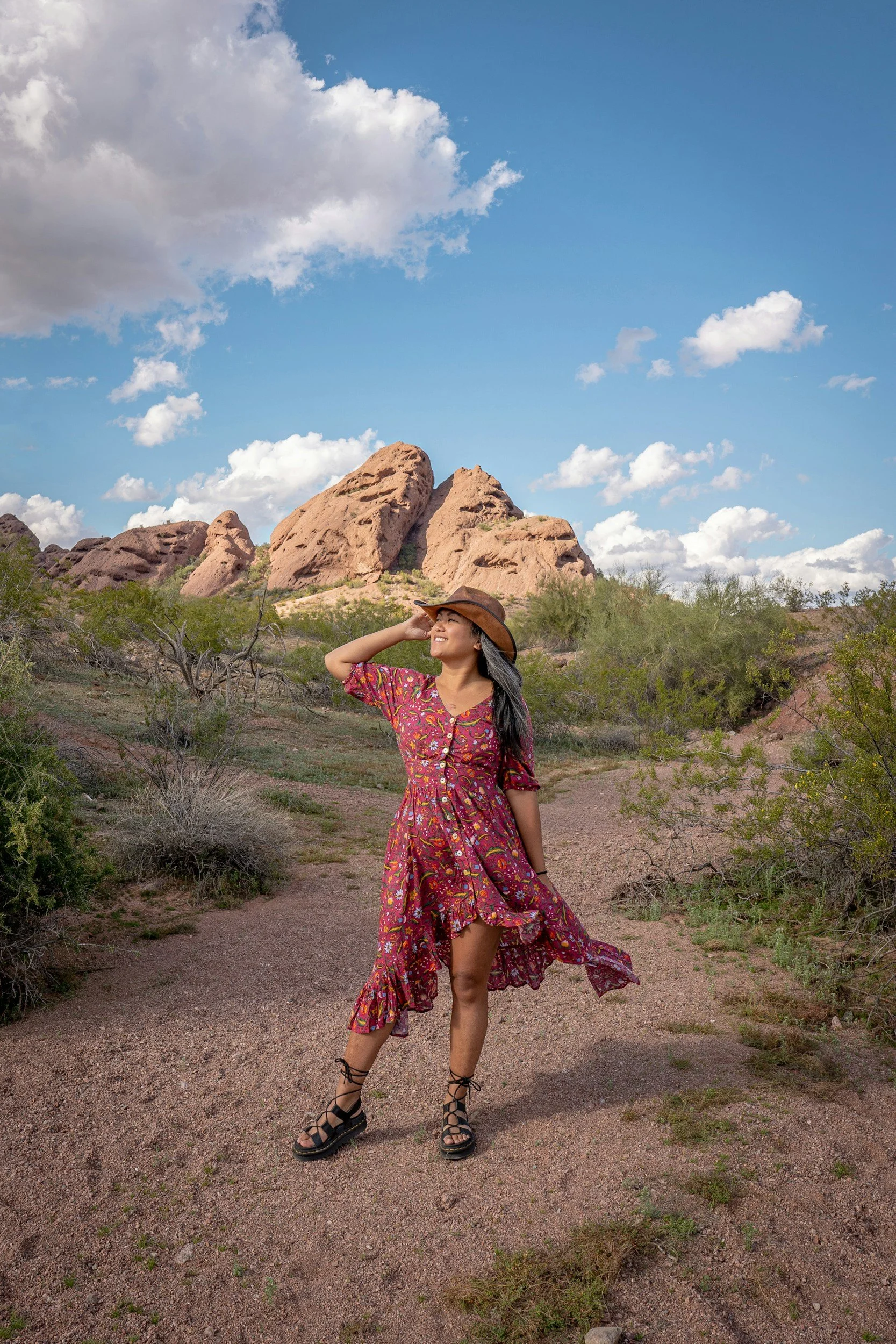 A woman in a floral dress, black gladiator sandals, and a wide-brimmed hat stands on a dirt trail in a desert landscape with large reddish rock formations and sparse desert vegetation under a blue sky with scattered clouds.