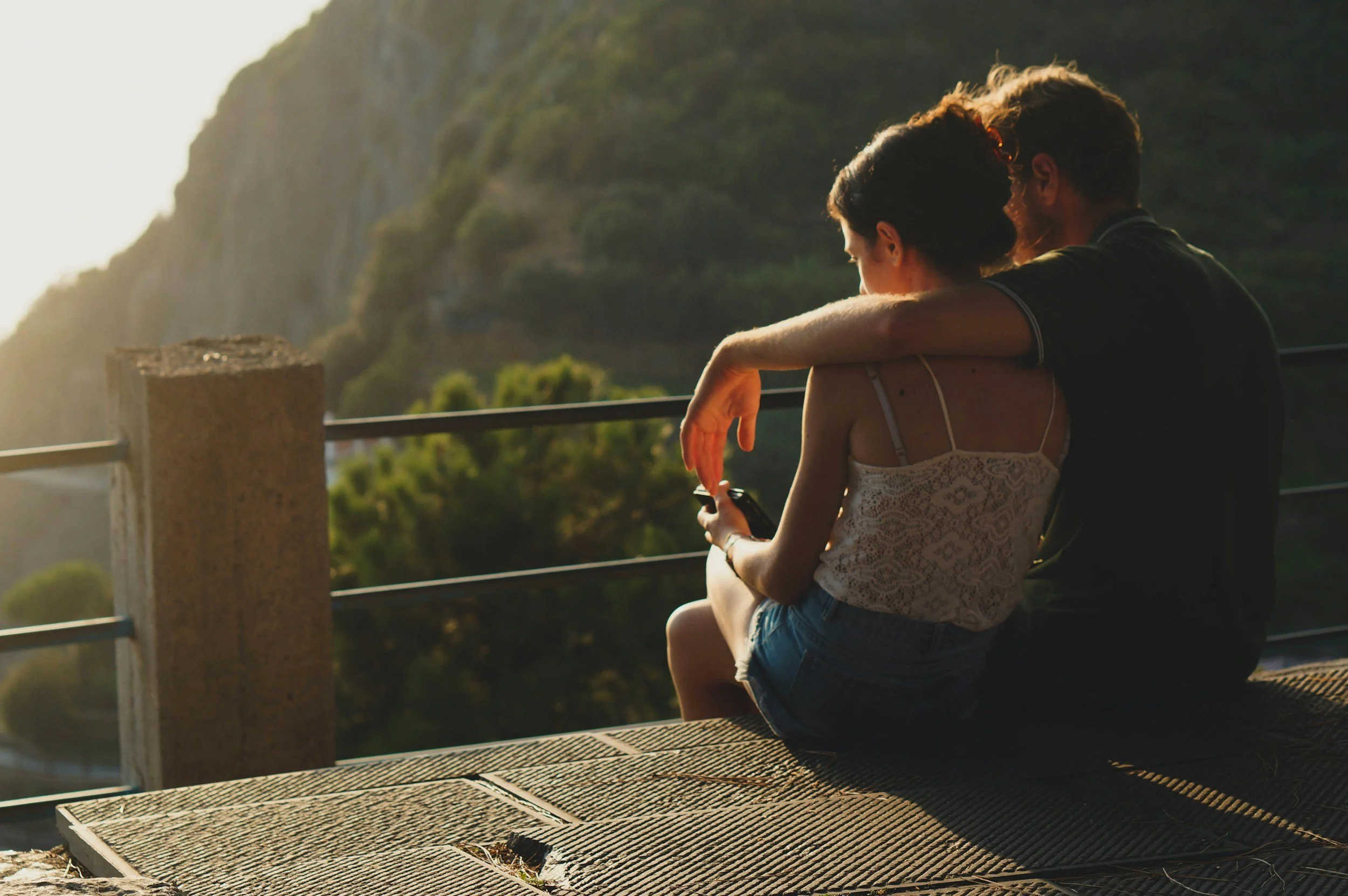 A young couple sitting on a wooden bench by a railing overlooking a scenic mountainous landscape during sunset, with the person in front holding a smartphone.