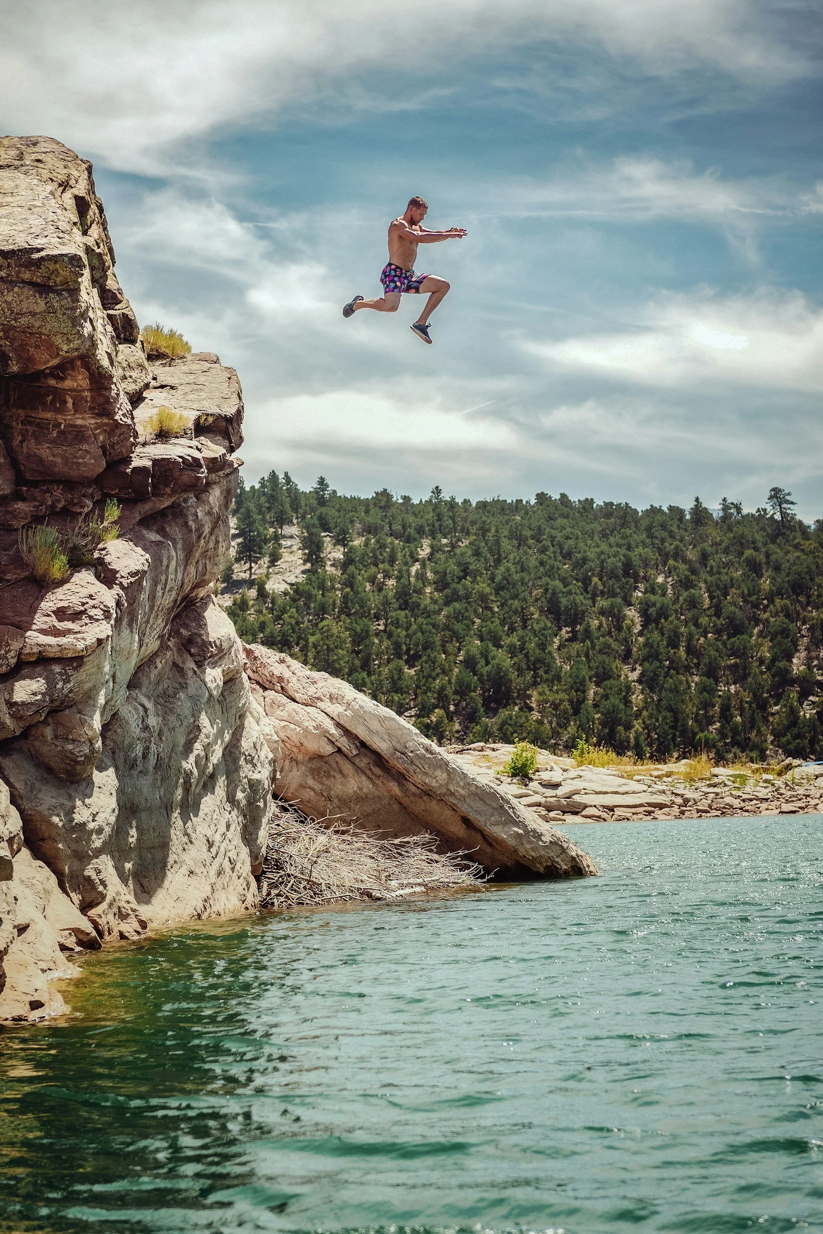 A man jumping from a rocky cliff into a body of water, with a backdrop of a forested landscape and a cloudy sky.