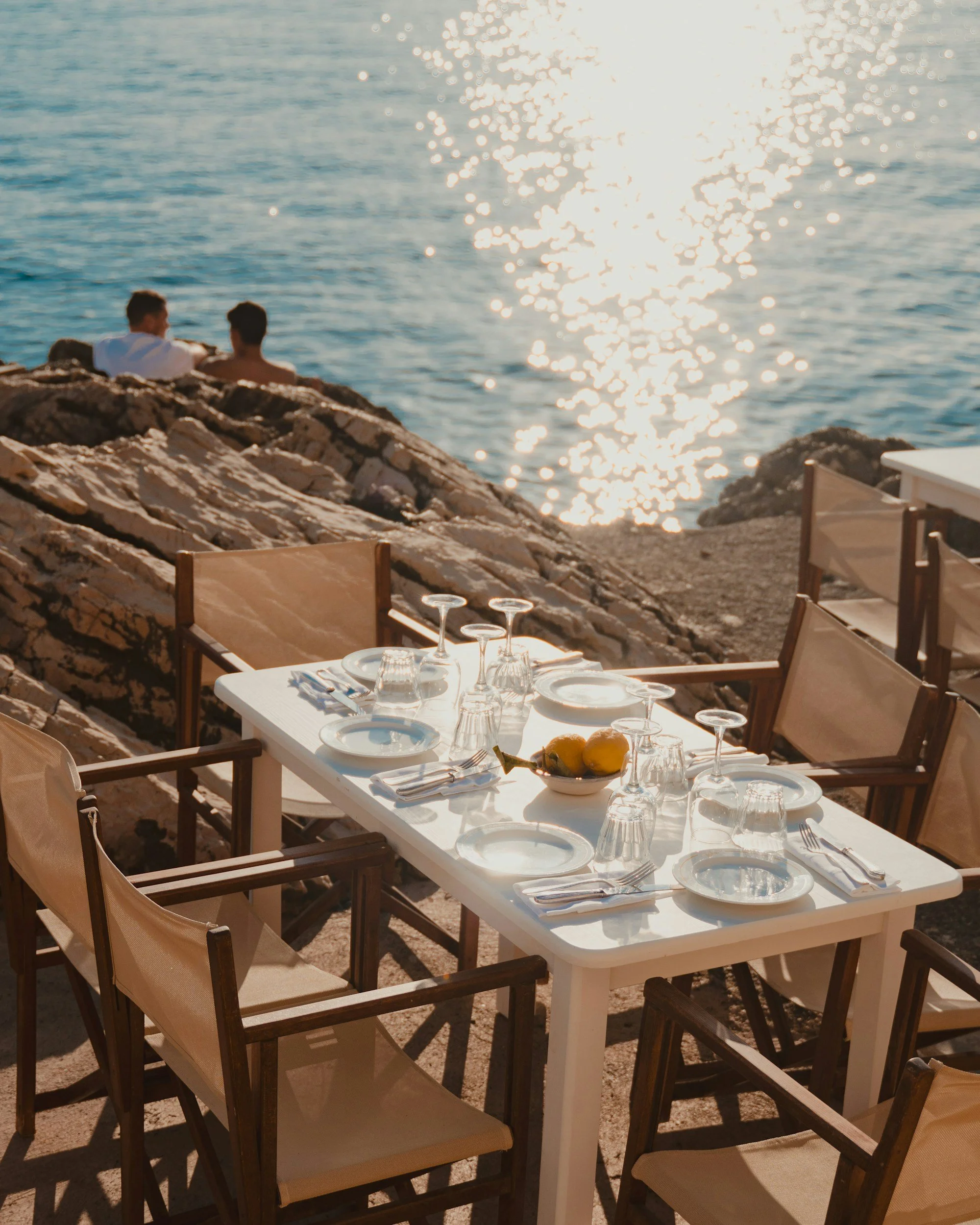 A white outdoor dining table set for a meal with glasses, plates, silverware, and a bowl of lemons on a rocky beach with sun reflecting on the water in the background.