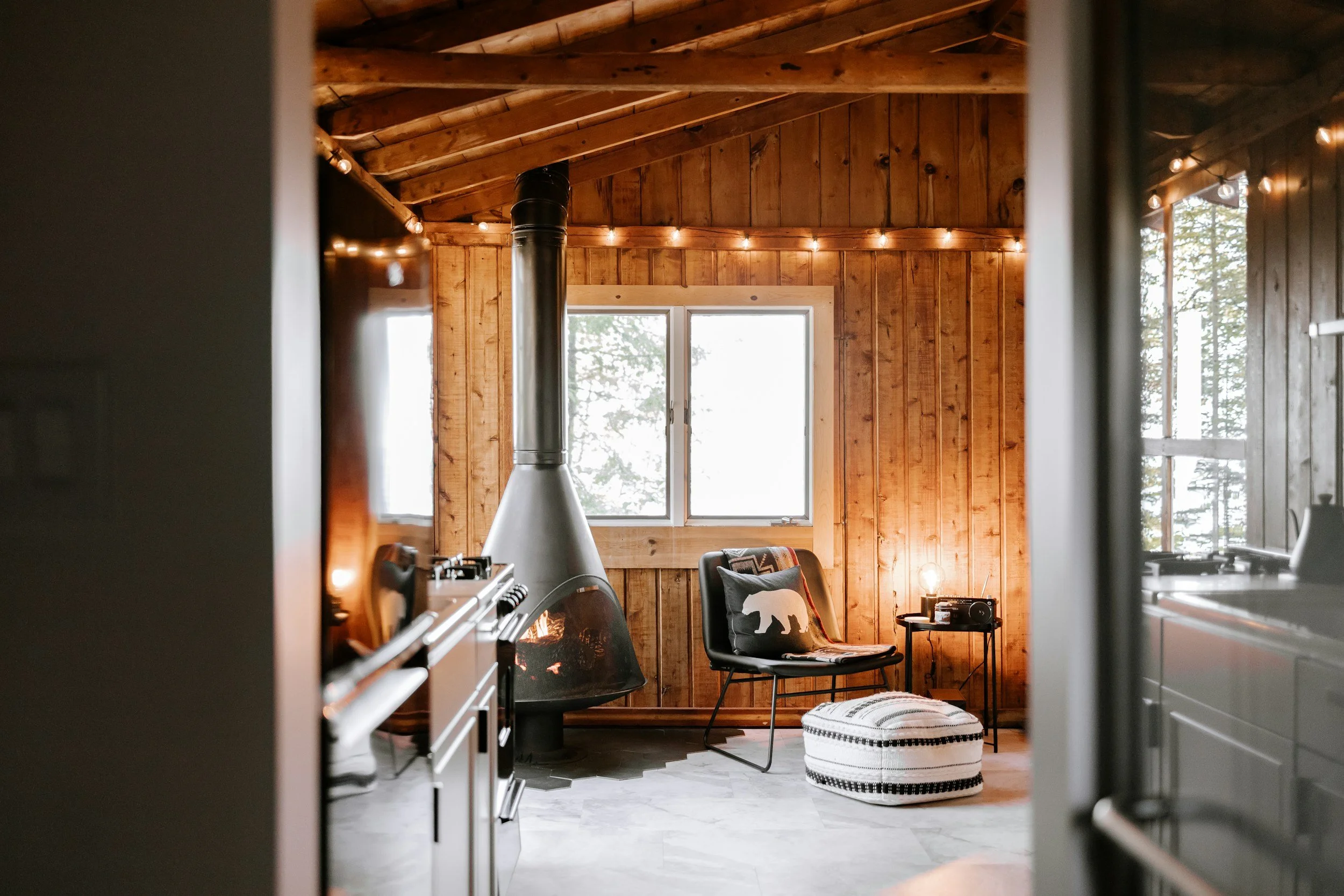 View from a kitchen into a cozy living space with wooden walls and ceiling, large windows, a black chair with a pillow, a small side table, a pouf at the foot of the chair, and string lights hanging across the ceiling.