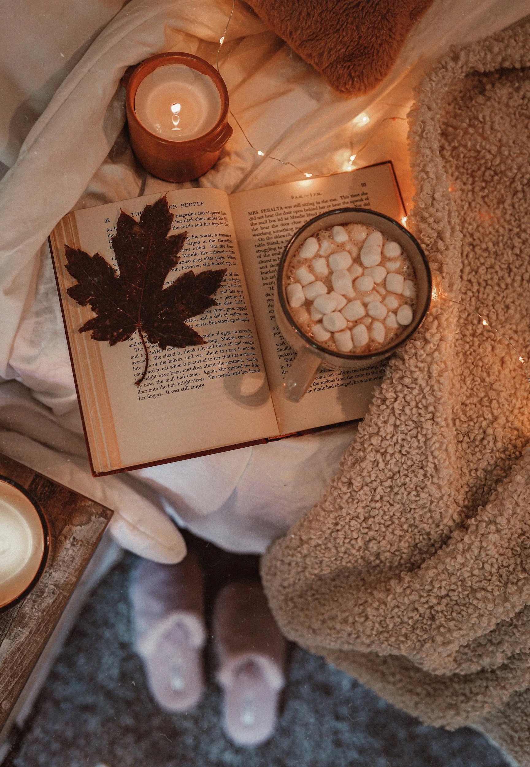 Open book on a table with a dried leaf on the page, a cup of hot chocolate with marshmallows, a candle in a brown mug, and string lights, surrounded by cozy blankets.