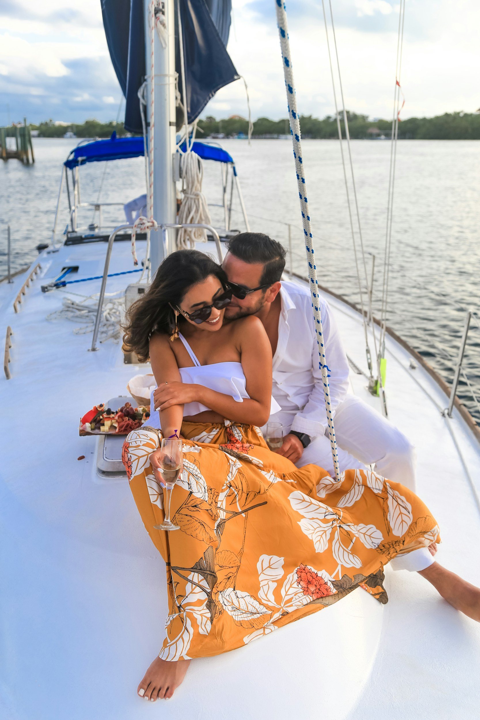 A couple on a boat, the woman wearing orange patterned skirt and sunglasses, holding a glass of wine, the man leaning into her, both smiling, with water and trees in the background.
