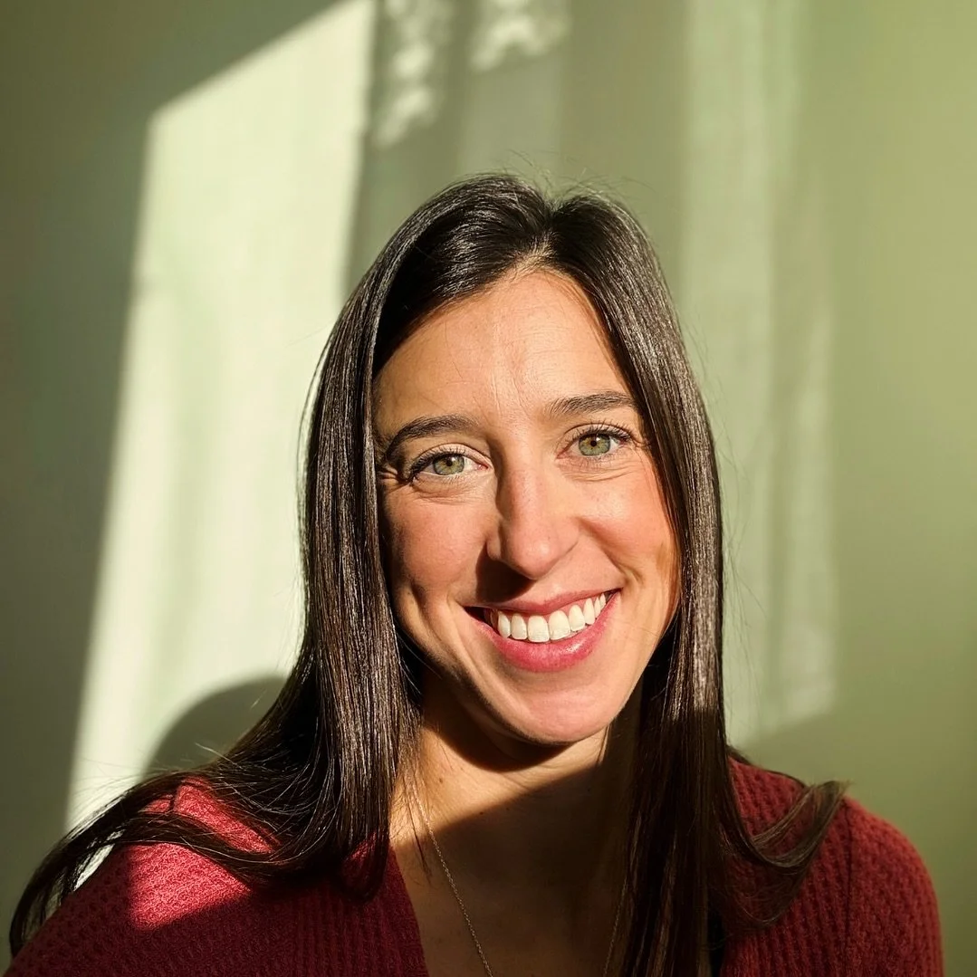 A woman with long dark hair and green eyes smiling, with sunlight casting shadows on a light-colored curtain behind her.