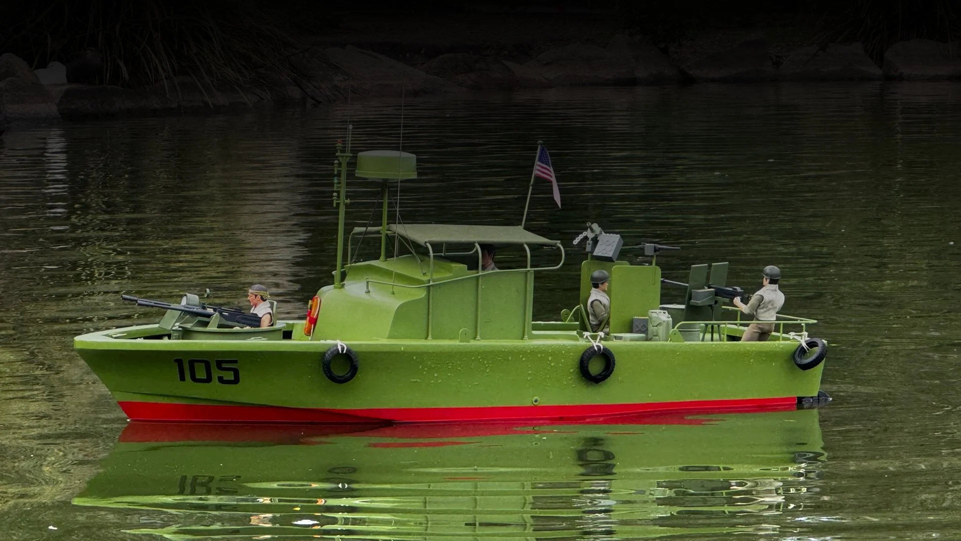 A toy military boat painted in bright green with the number 105 on the side, floating on water. It has four toy soldiers on board, some aiming guns and others operating equipment. The boat features an American flag, antennae, and life rings hanging on the side.