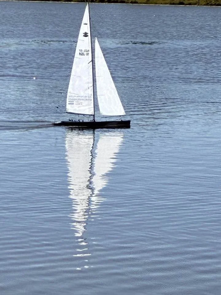 A small model sailboat floating on calm water with its reflection visible.