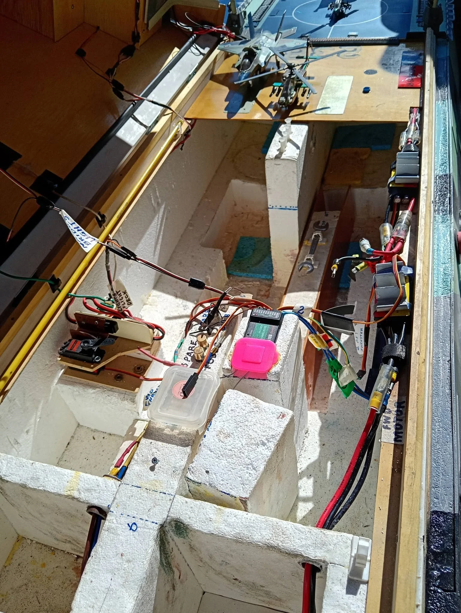 A workbench with model helicopters, electrical wires, tools, and equipment, set in a workshop or garage environment.