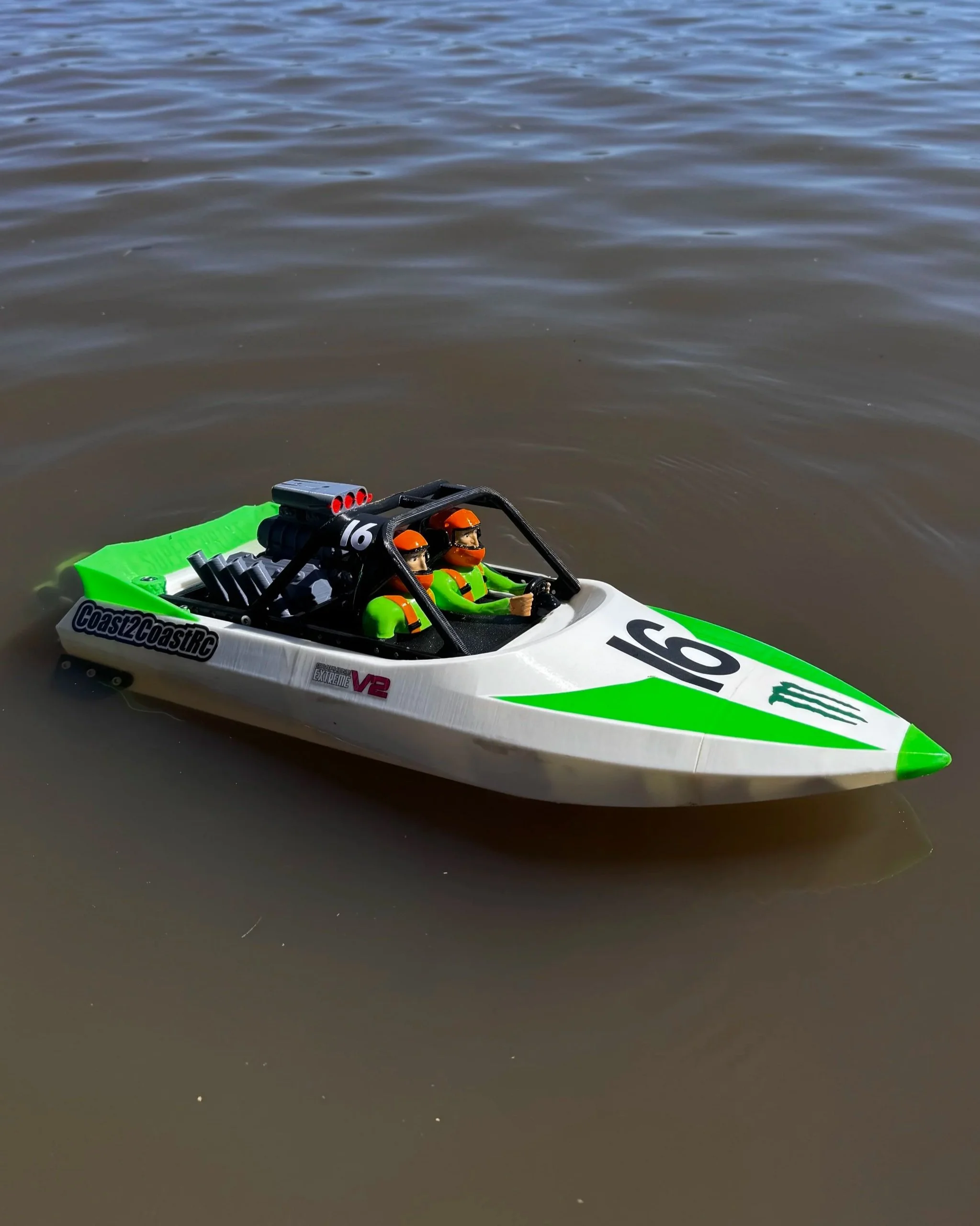 A remote-controlled speedboat with two toy figure pilots racing on a calm, brown water surface.