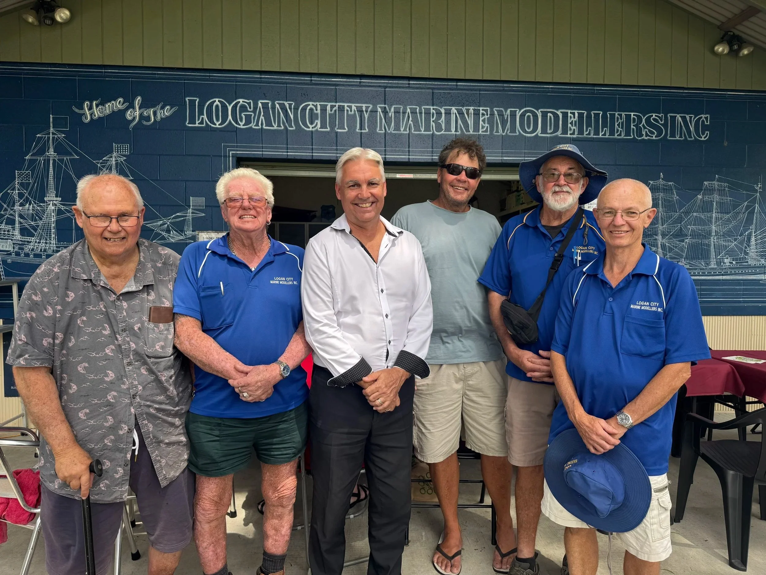 Group of six men standing together in front of a mural at the Logan City Marine Modellers Inc. building; some wearing sunglasses and casual clothes, smiling for the camera.