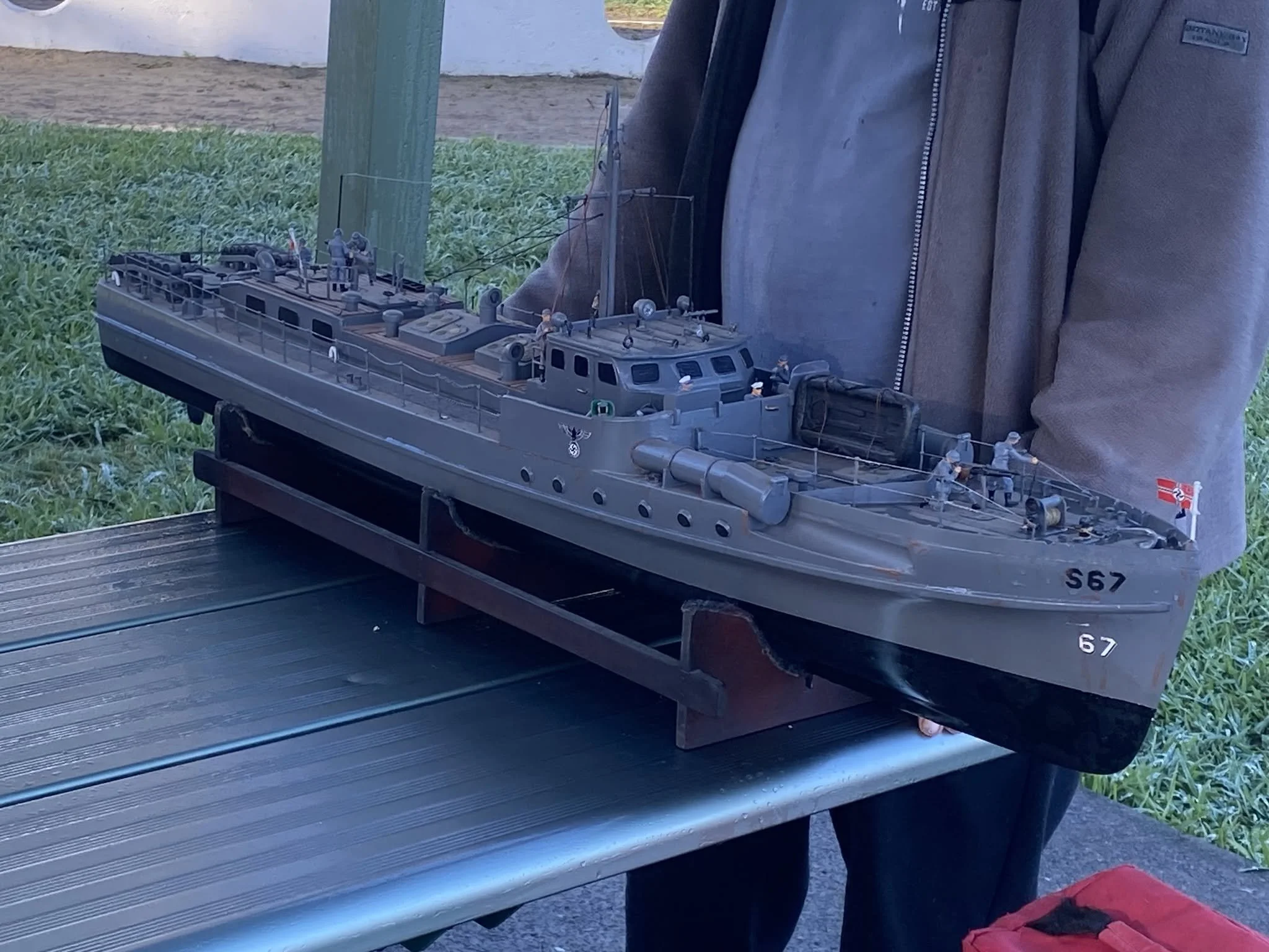 A person wearing a gray zip-up jacket standing next to a detailed scale model of a battleship displayed on a black surface outdoors.