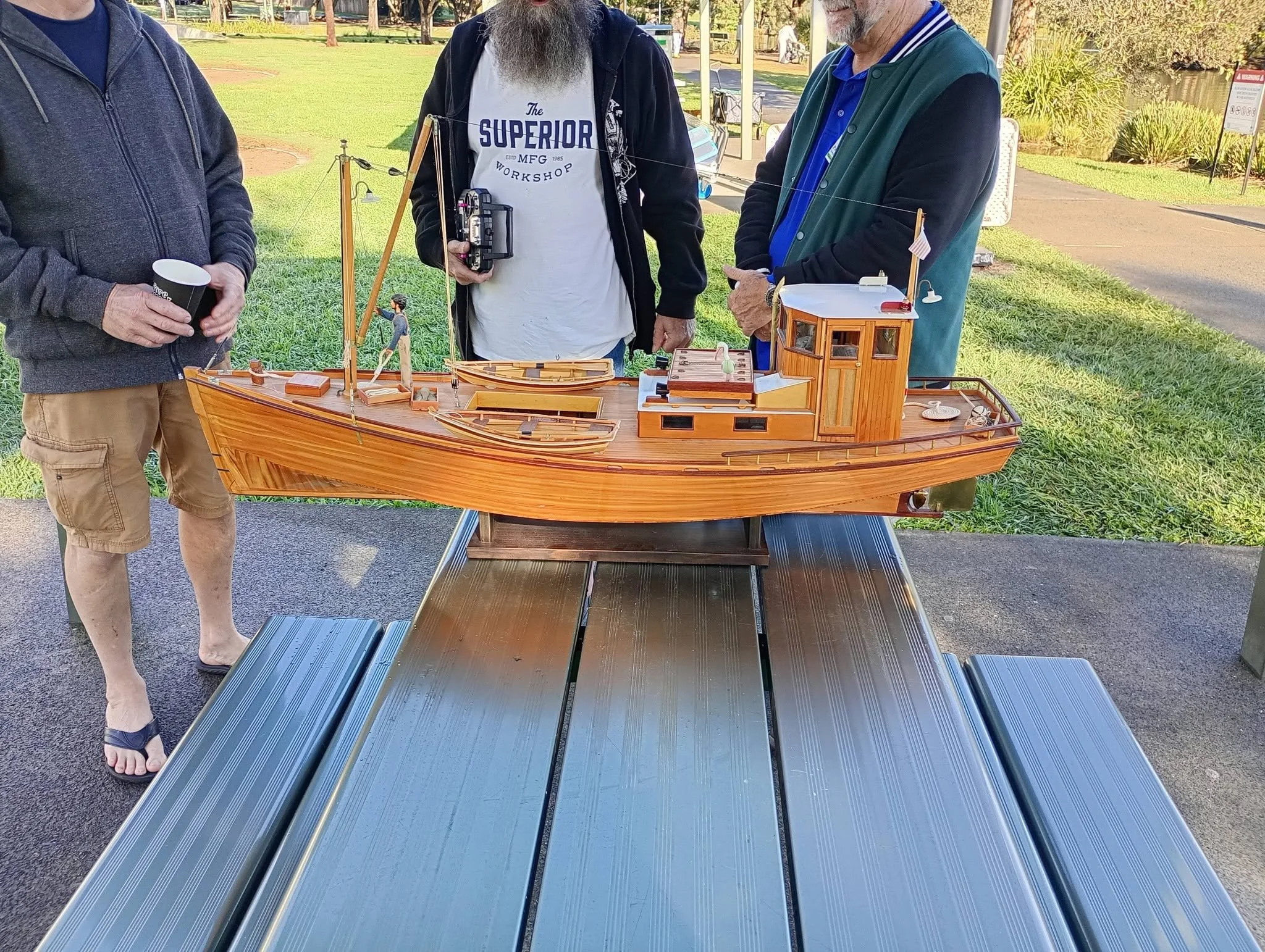 Three men are standing around a model sailing ship displayed on a table outdoors in a park. One man is holding a paper cup, another has a remote control, and the third has his hands clasped. The ship is detailed with small figures, buildings, and dec