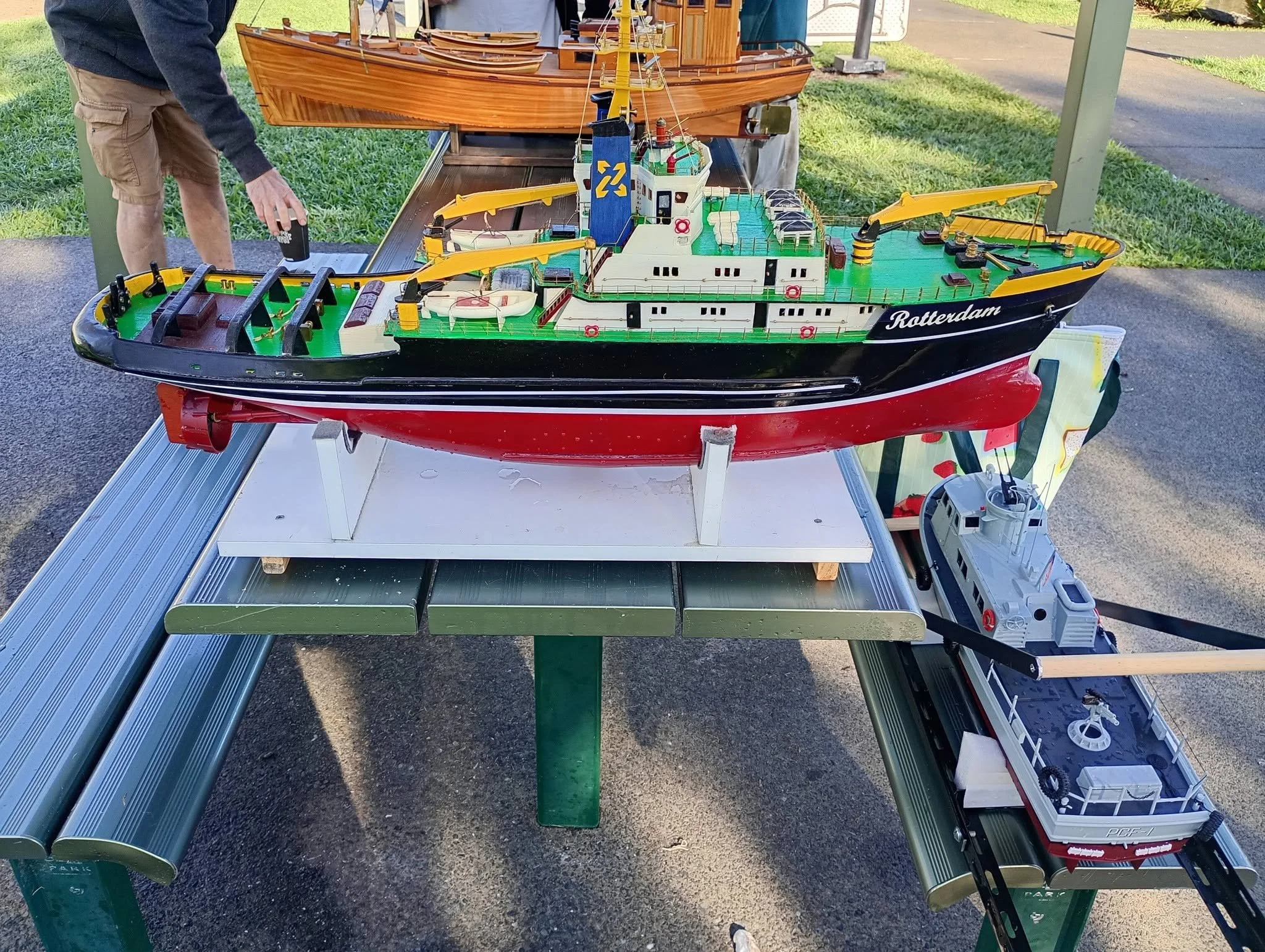 A detailed model of a large ship labeled 'Rotterdam' on its side, displayed outdoors on a table with various tools and equipment nearby, and people observing it.