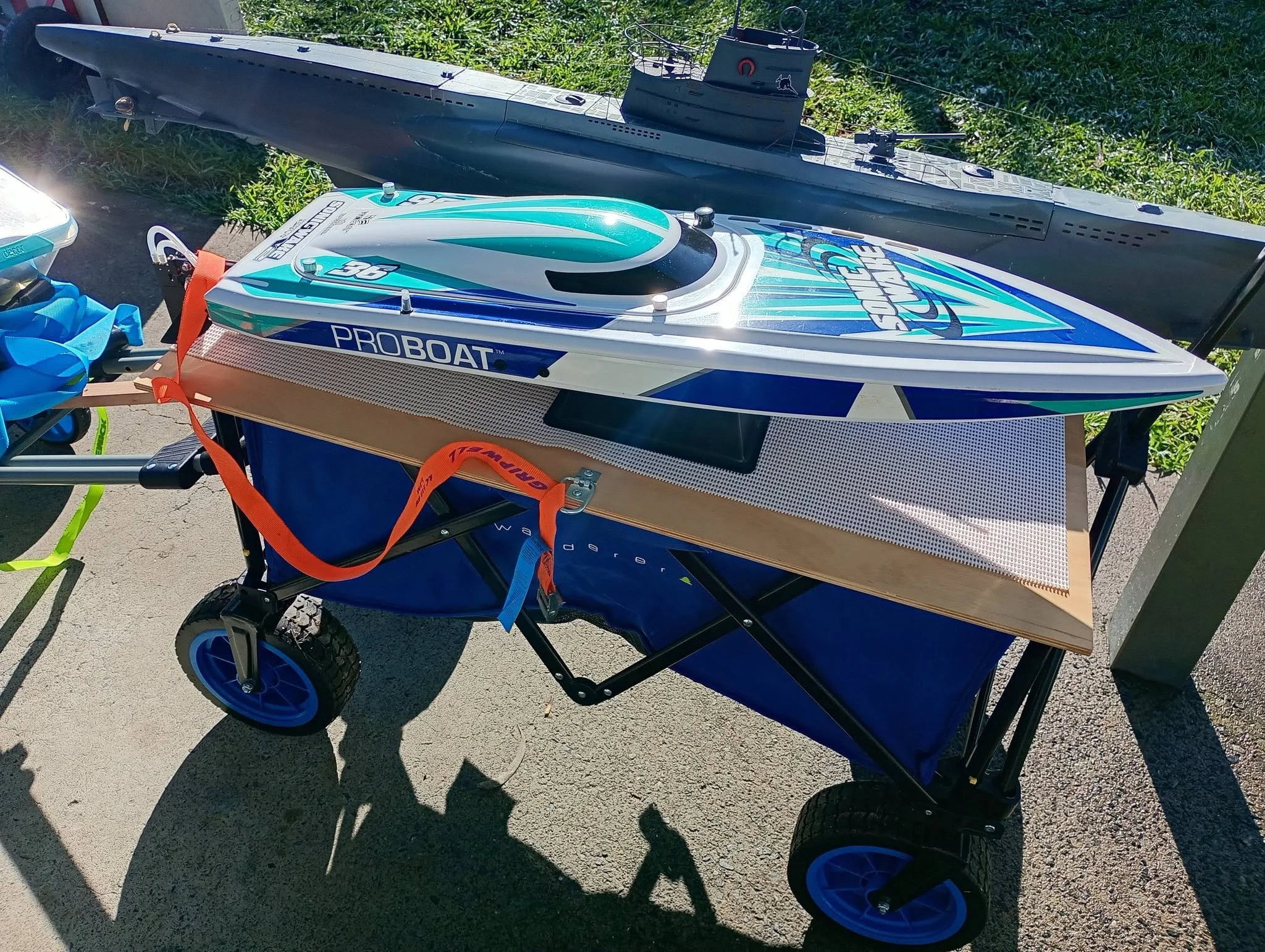 A model boat on a cart with blue wheels, placed outdoors on a concrete surface with grass in the background.