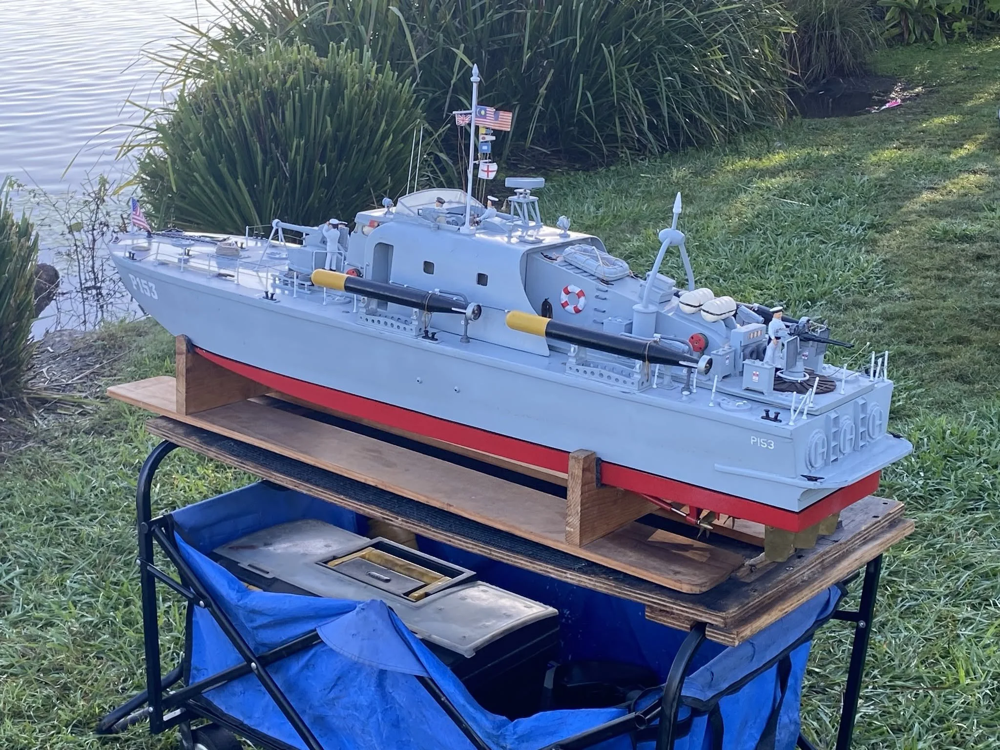 A detailed model of a gray military ship with small figures of soldiers on board, placed outdoors on a wooden table near water and tall grass, with flags and equipment visible on the ship.