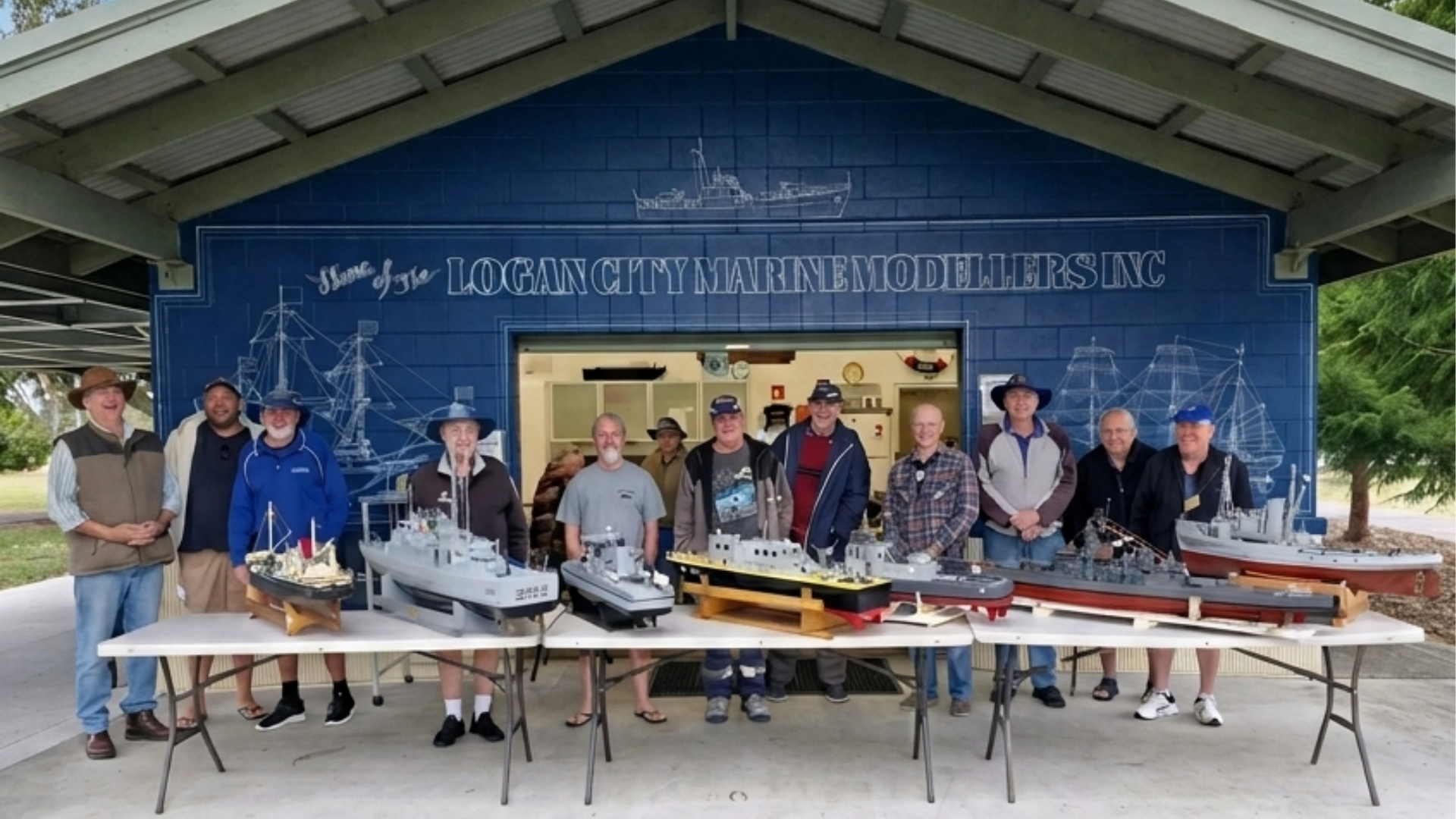 Group of mostly men standing behind tables with model ships at the Logan City Marine Modelers Inc. clubhouse, with a blue sign overhead.