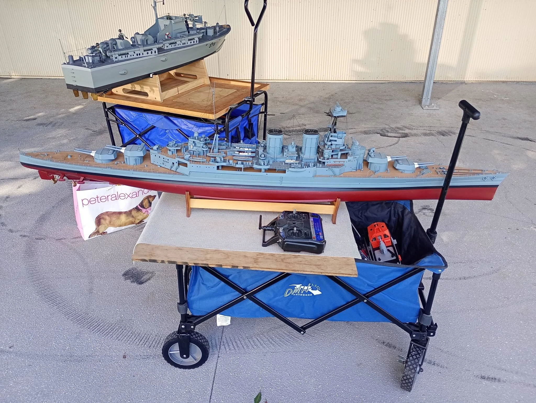 A display of detailed model ships on a table and a stand outdoors. The larger ship model is placed on a wooden stand, and nearby are smaller ship models. The setup includes a black and red remote control and a blue outdoor chair with a pink and white