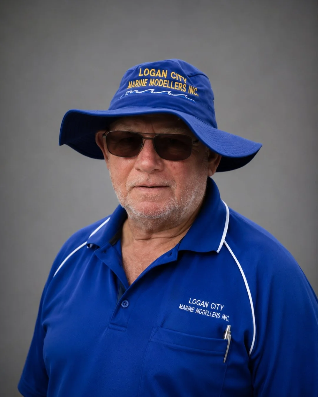 Older man wearing a blue wide-brimmed hat with yellow embroidery that reads 'Logan City Marine Modellers Inc' and a logo of waves. He has on dark sunglasses and a blue polo shirt with white piping, also embroidered with 'Logan City Marine Modellers Inc.' smiling at the camera against a plain gray background.