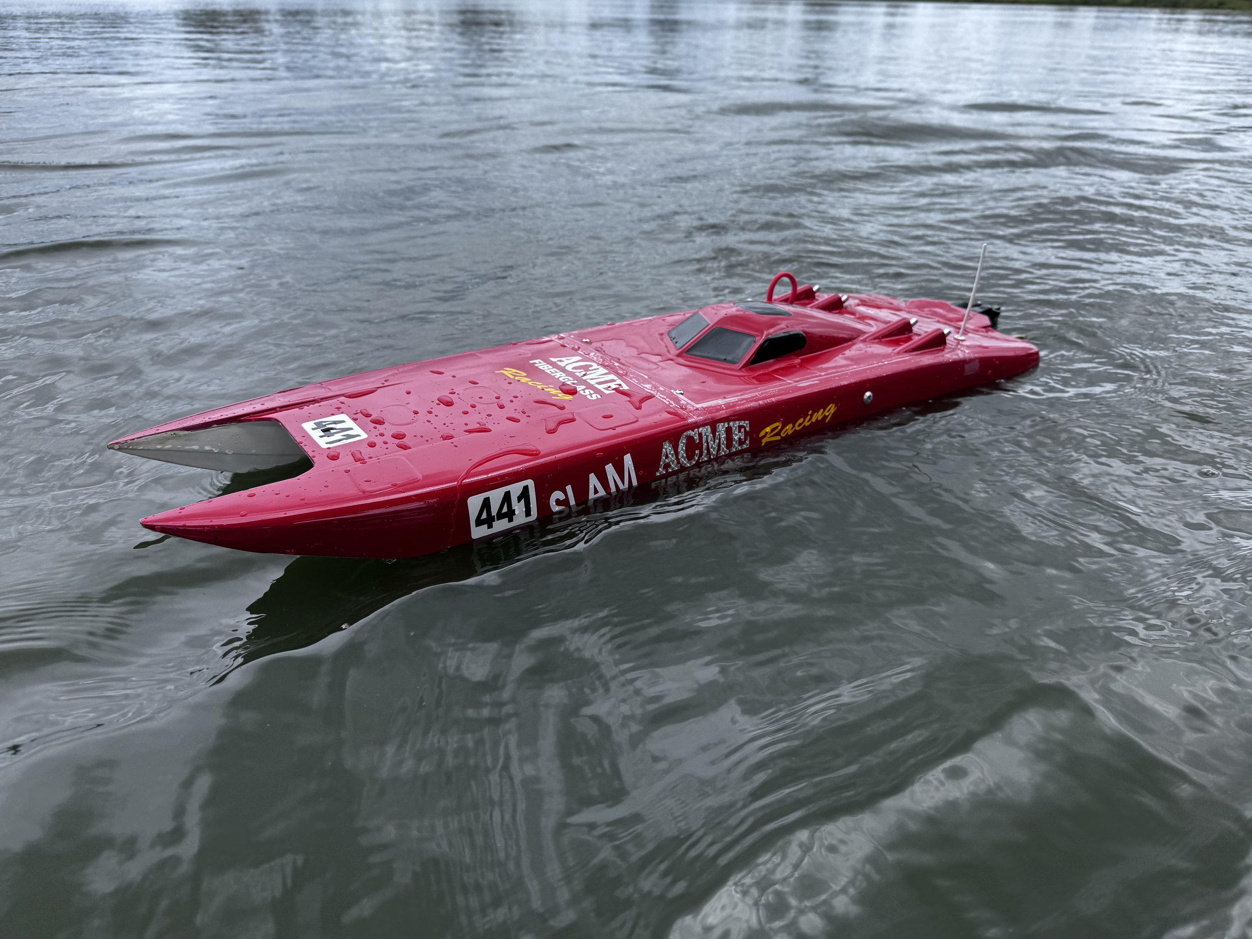 A red remote-controlled speedboat floating on water with small ripples, water droplets on the boat, and various sponsor logos including 'ACME Racing' and 'SLAM'.