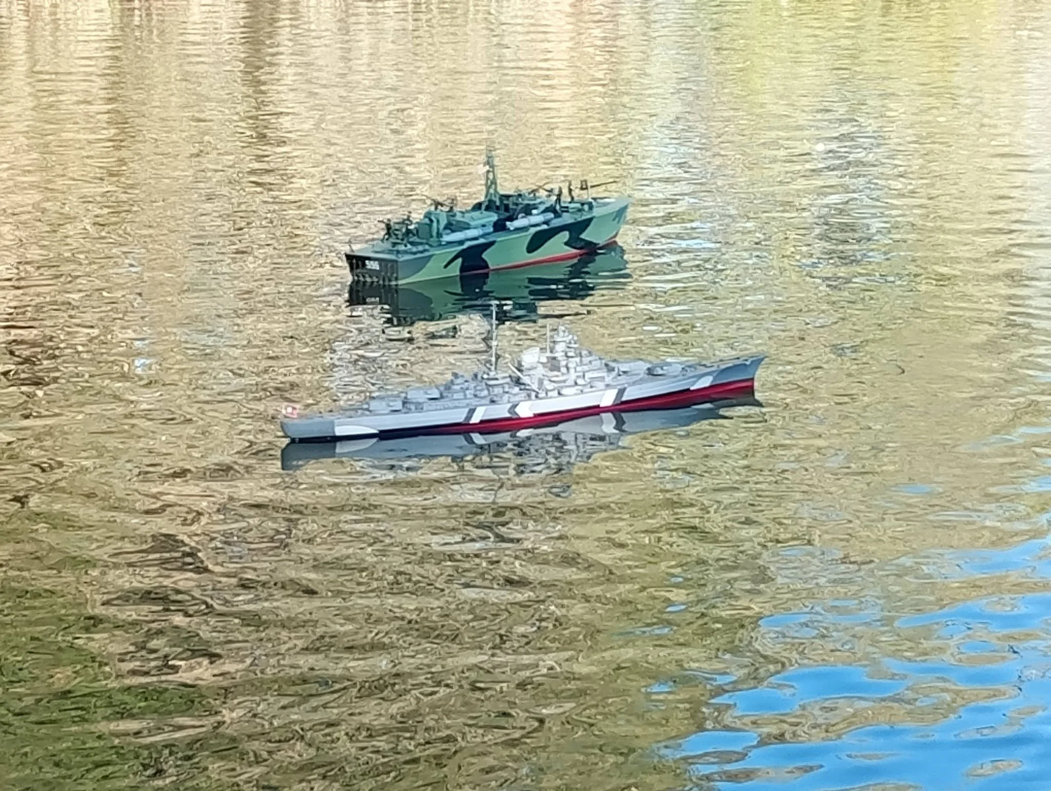 A small model ship with a green, black, and red camouflage pattern on the water, with reflections of the ship visible on the water's surface.