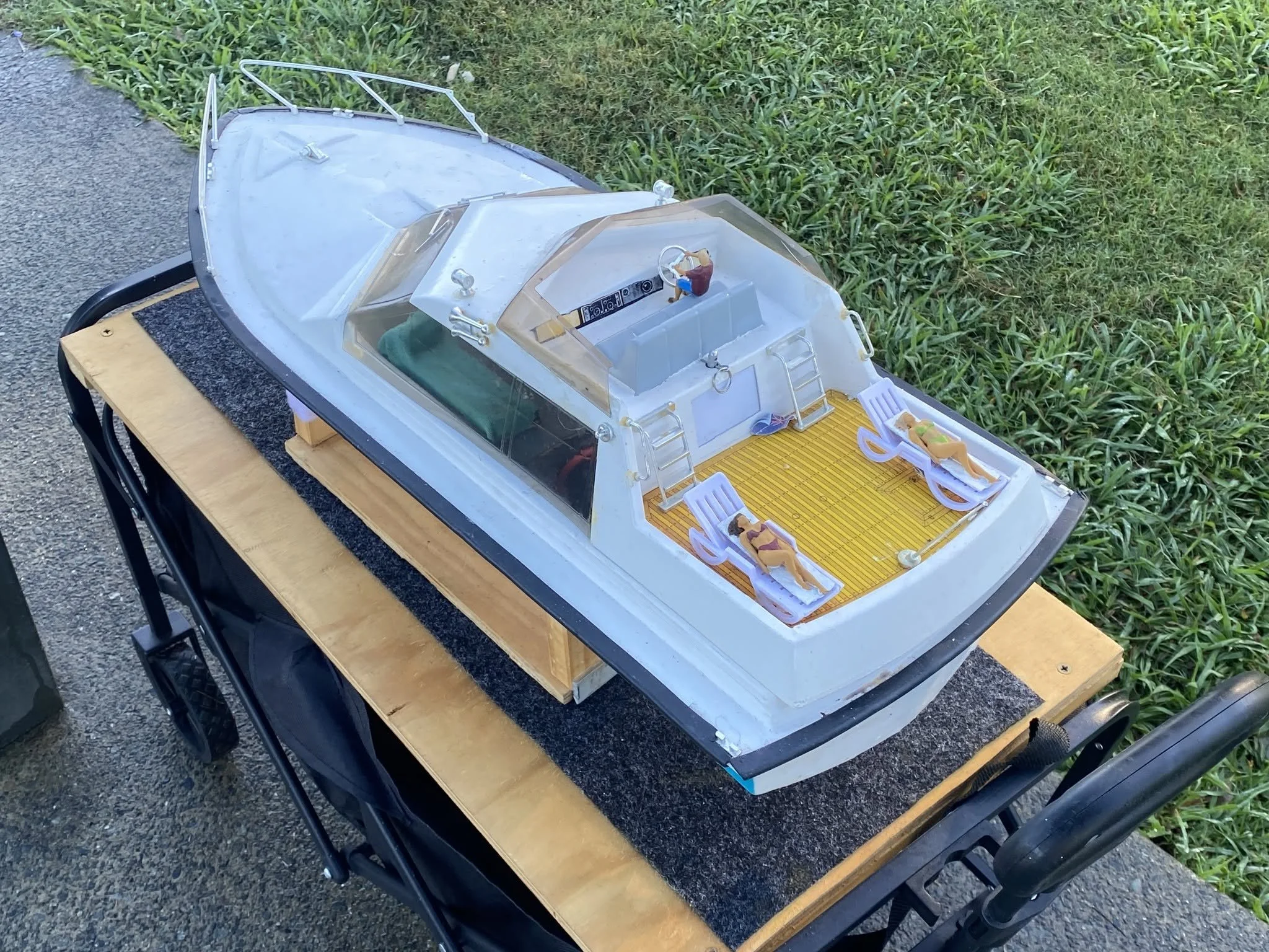 Model of a boat with a clear plastic cover, small figurine in driver's seat, and miniature chairs, placed on a wooden table outdoors.