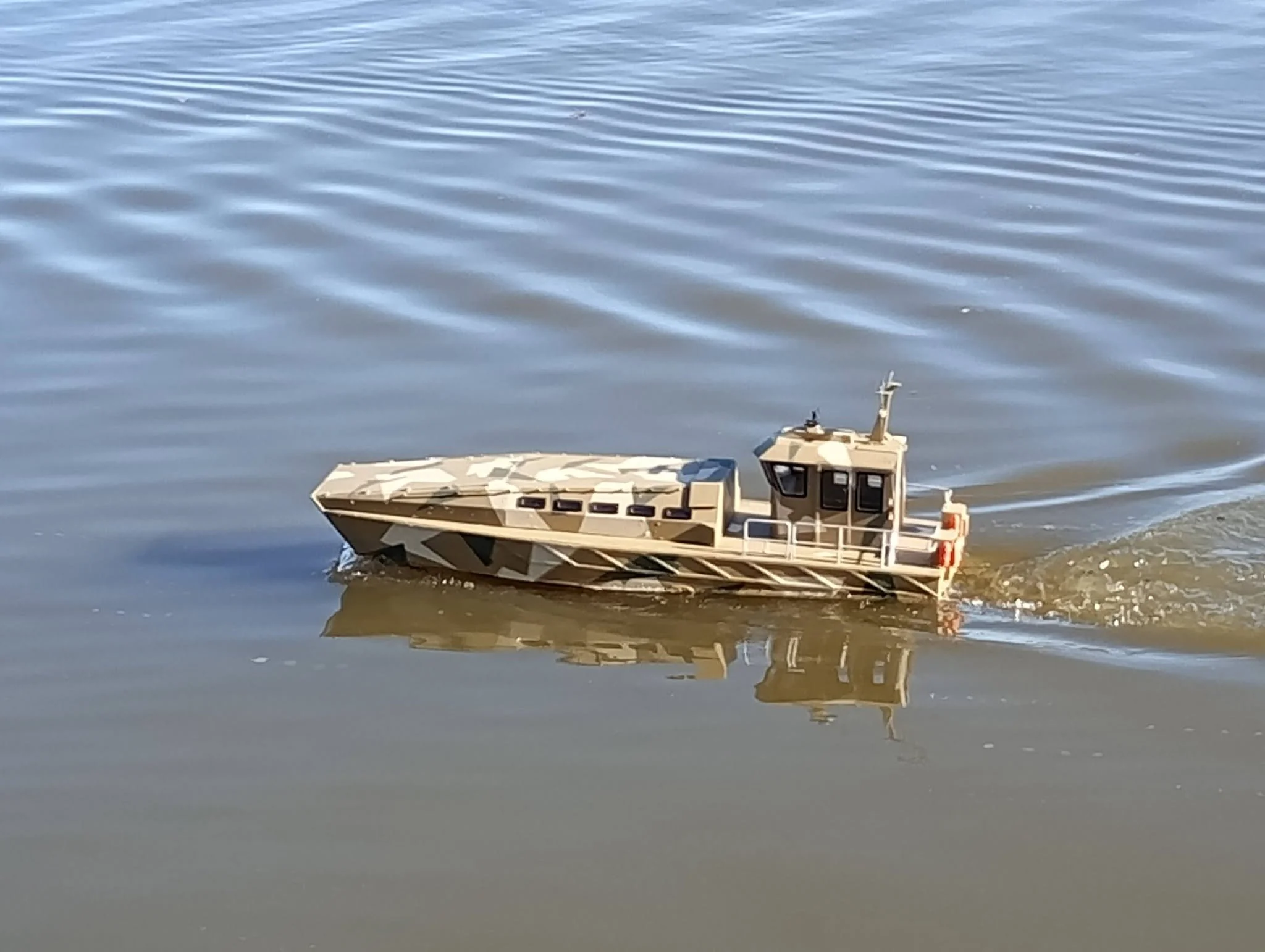 A military-style camouflage boat floating on water with visible ripples. Reflection of the boat can be seen on the water surface.