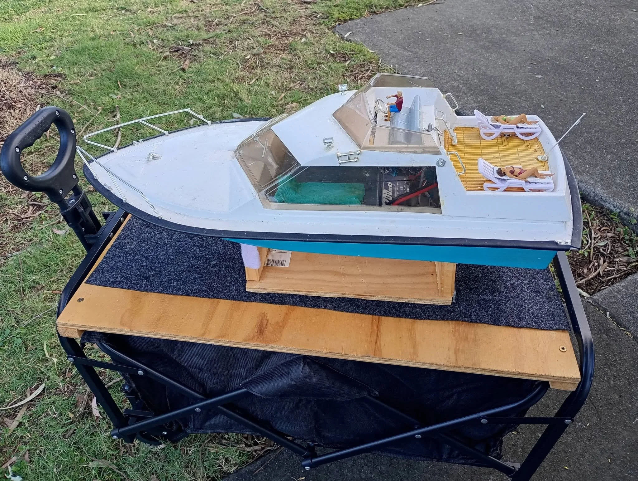 A remote-controlled boat with a white and blue hull, a clear cabin, and miniature figurines on board, sitting on a wooden table outdoors next to grass and pavement.
