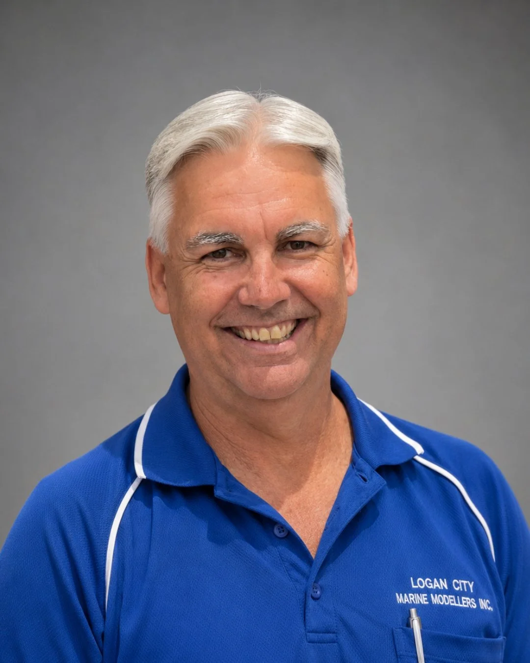 A smiling middle-aged man with gray hair wearing a blue polo shirt with white piping, embroidered with 'Logan City Marine Modellers Inc.' against a gray background.