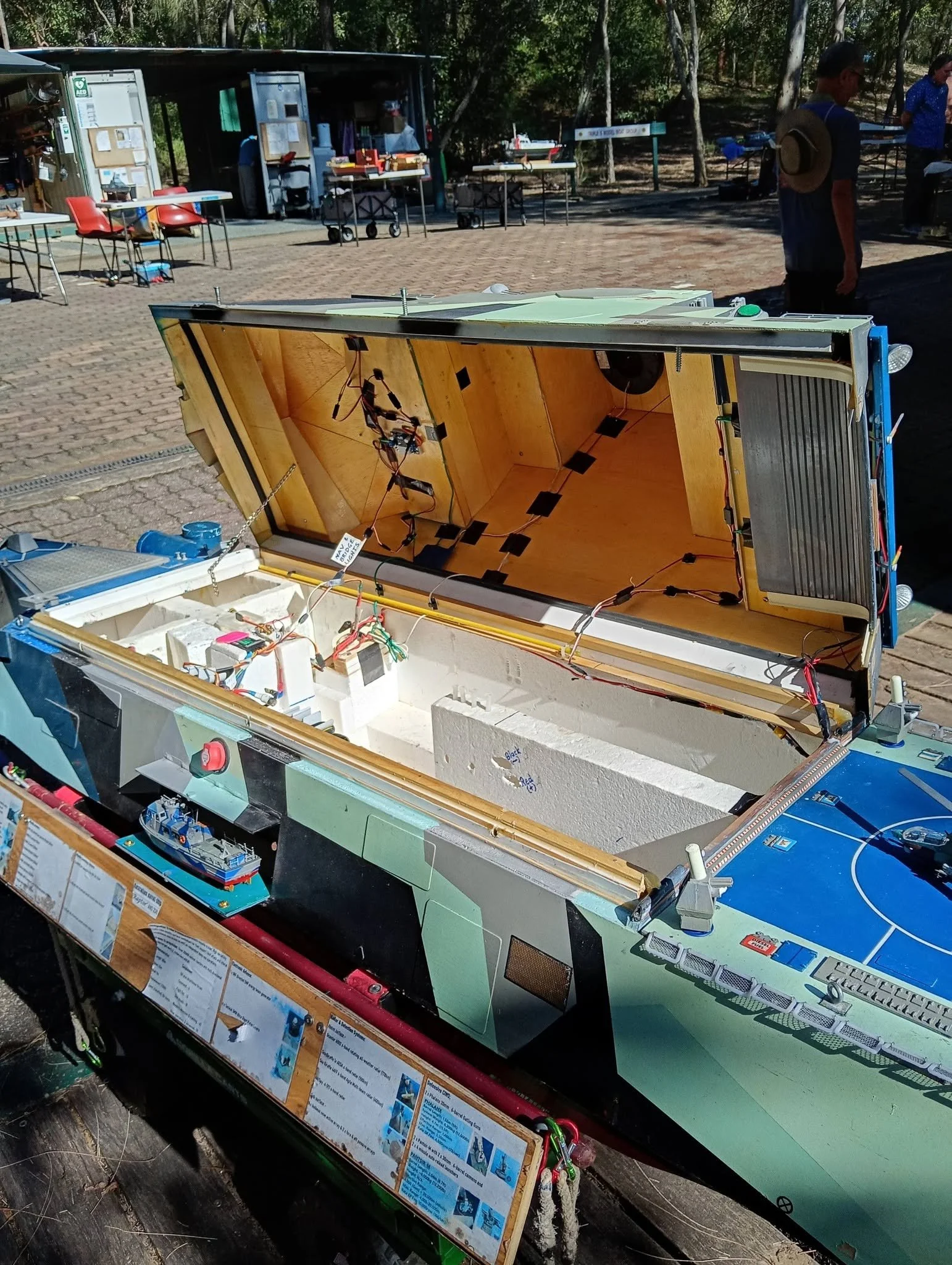 A model boat or ship under construction or display, with internal wiring and components visible inside the hull, at an outdoor setting with tables and people in the background.
