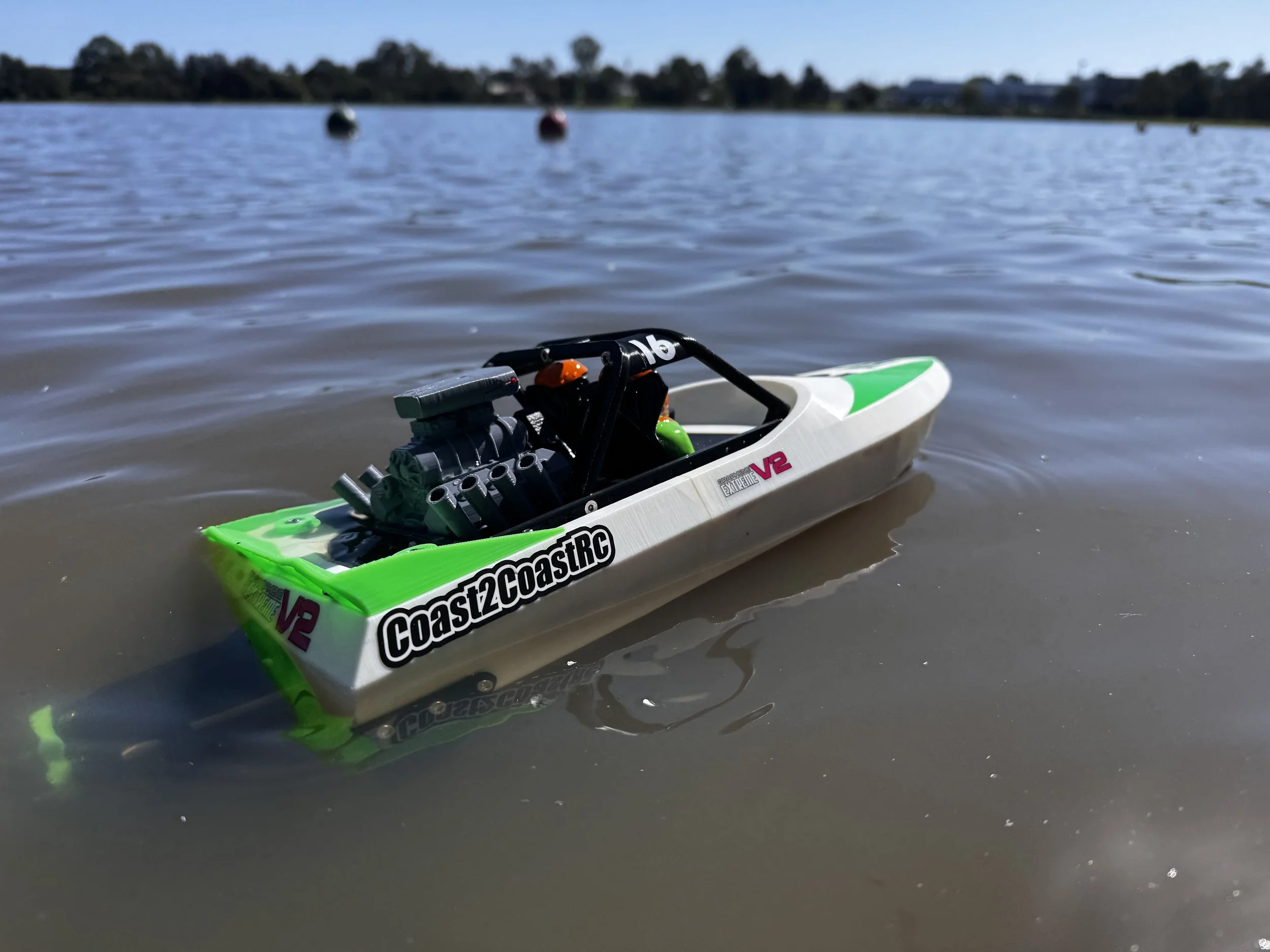 A remote-controlled boat floating on calm lake water, with trees and houses in the background.