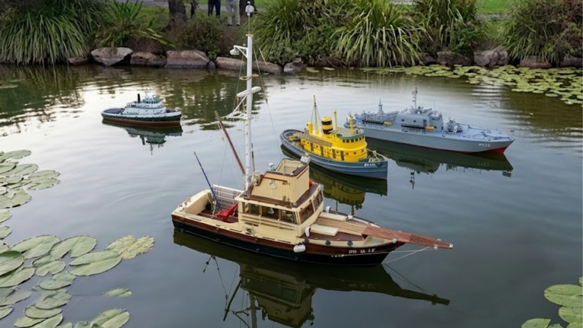 Four model boats floating on a pond with lily pads and rocks along the shore. In the background, a few people are visible standing among greenery.