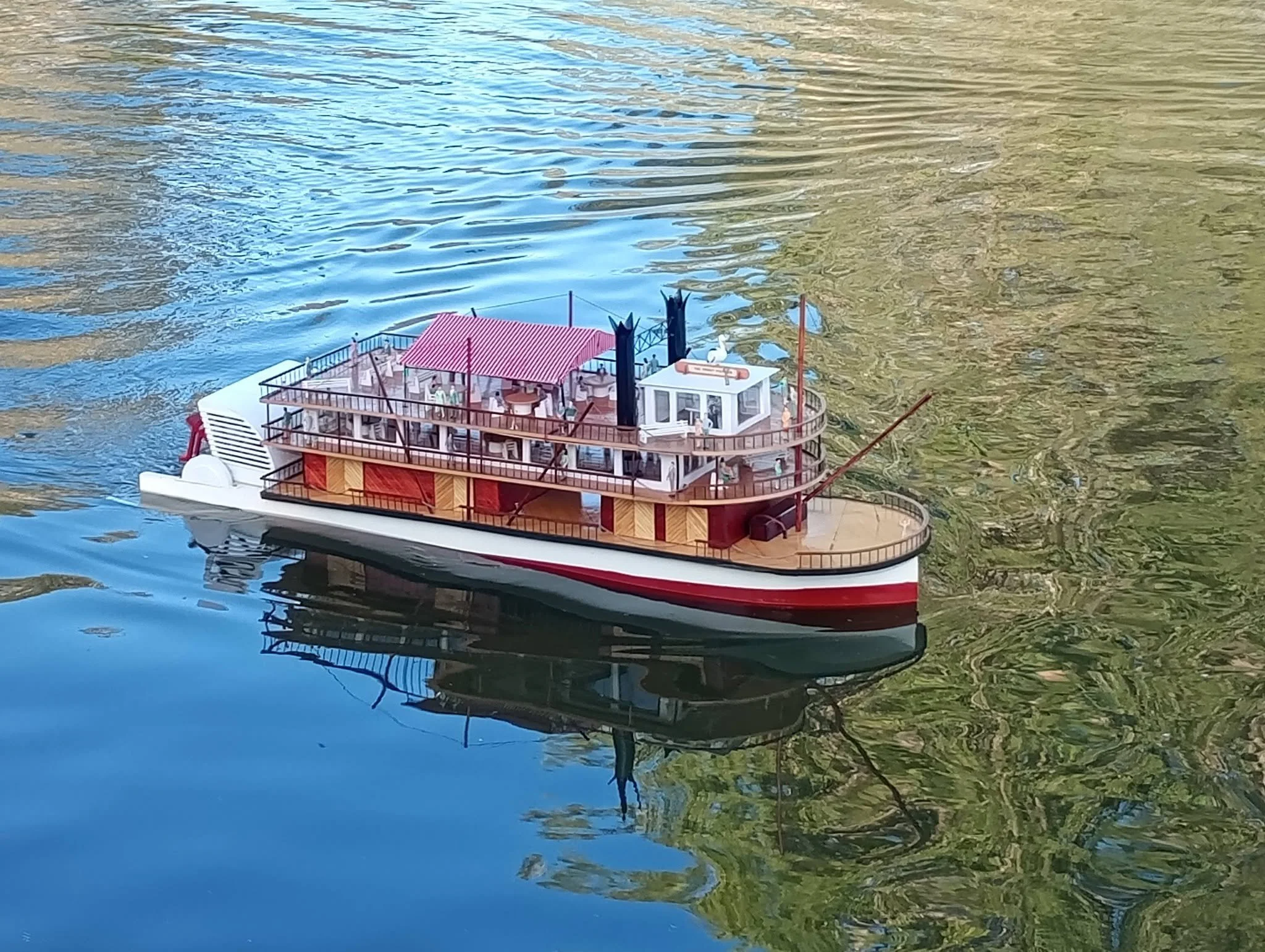 A detailed miniature model of a boat on water, featuring a red and white striped roof, blue smoke stacks, and tiny figurines, reflected in the water with surrounding green foliage.