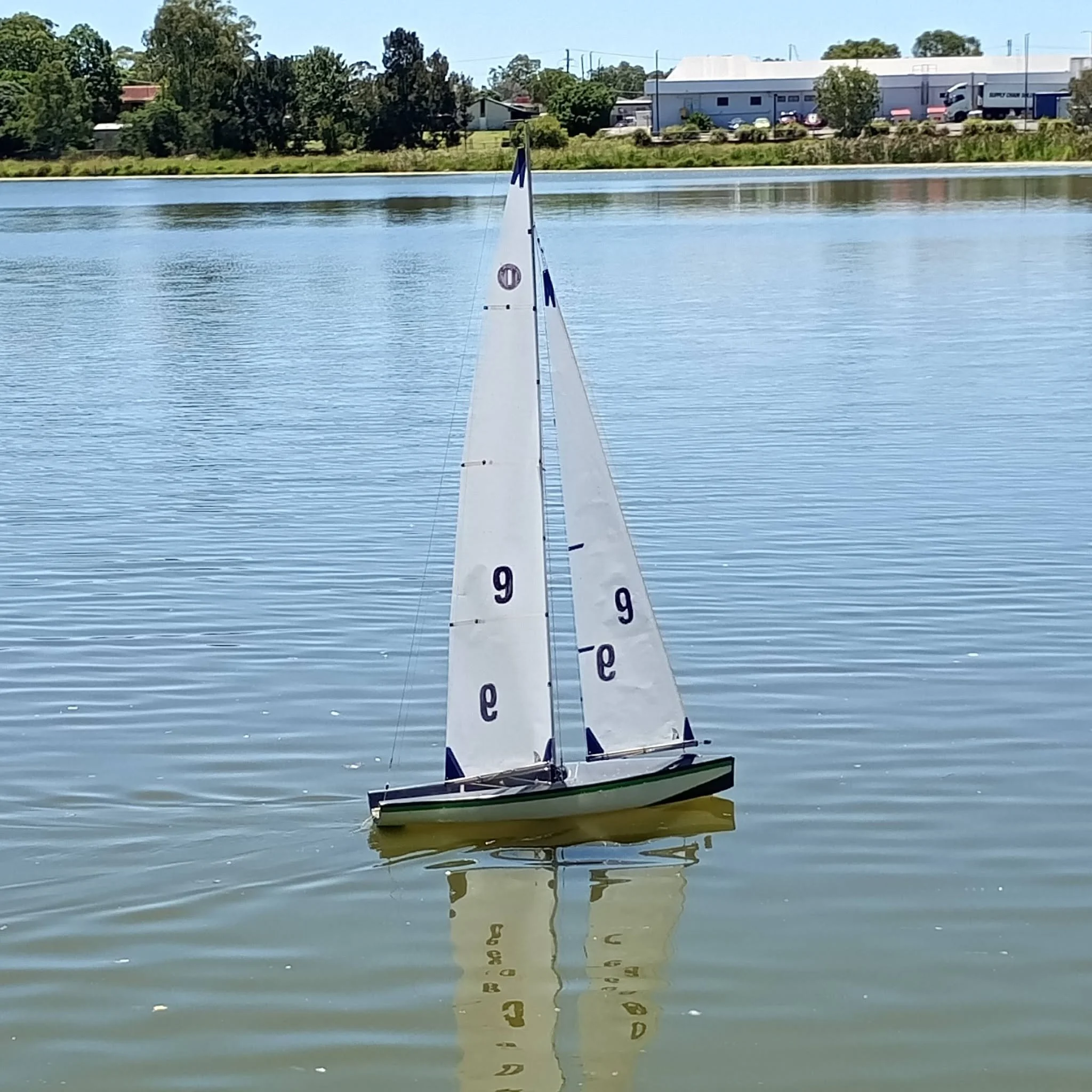 A model sailboat with the number 9 on its sail glides across a calm body of water, with trees and industrial buildings visible in the background on a clear, sunny day.