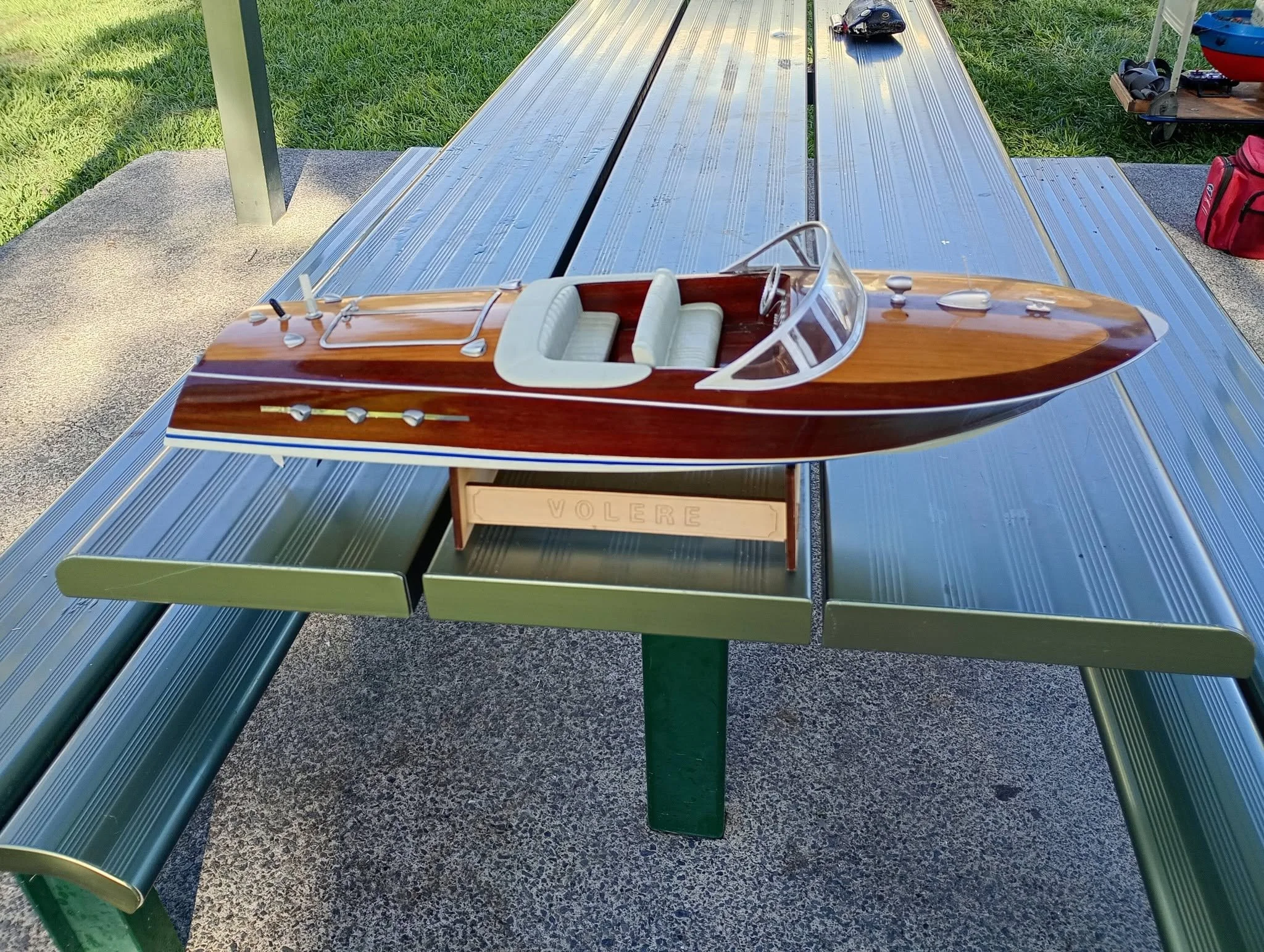A small wooden model boat with white seats and a windshield, placed on a metal table outdoors, with grass in the background.