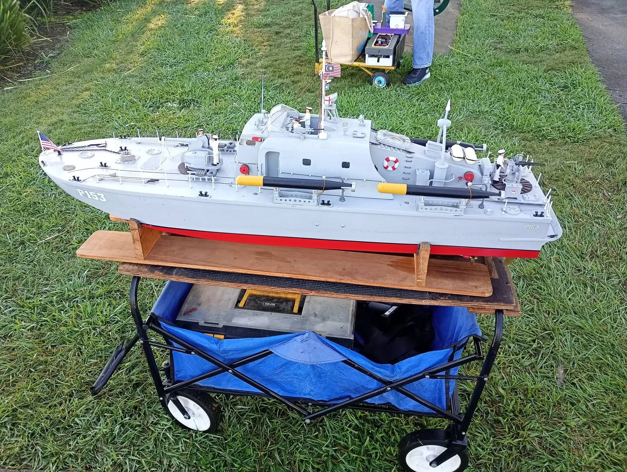A detailed model of a military battleship displayed outdoors on a wooden stand, with toy soldiers on deck, flags, and various mechanical features, next to a shopping cart with items.
