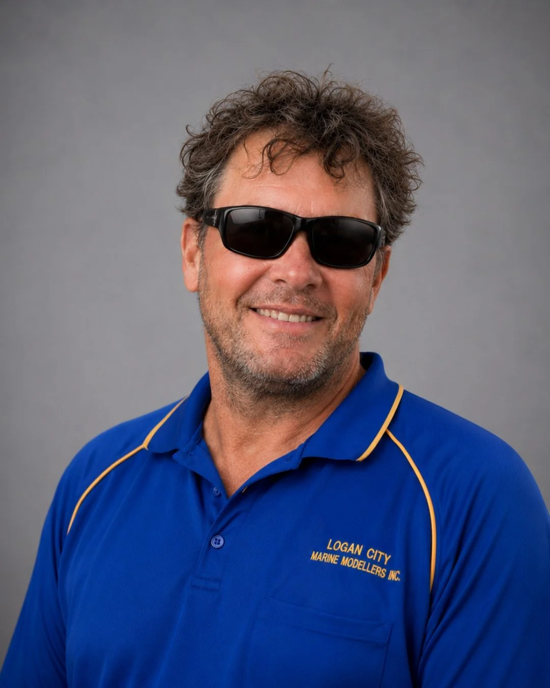 A middle-aged man with curly hair, beard, and sunglasses smiling in front of a gray background. He is wearing a blue shirt with yellow accents and the logo for 'Logan City Marine Modellers Inc.'