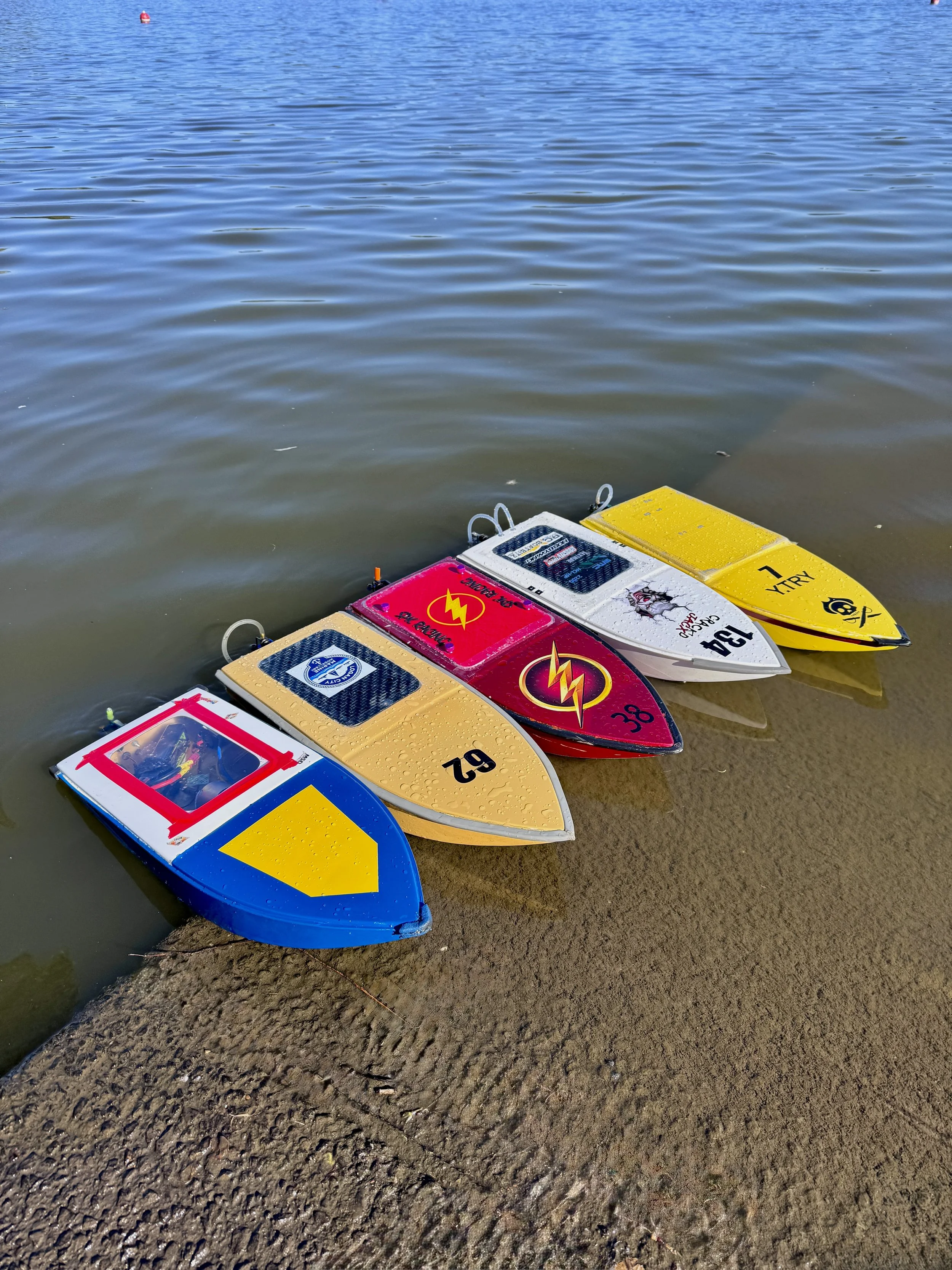 Four remote-controlled boats with colorful designs are lined up at the shore of a lake, partially in the water, with ripples on the lake surface and a small red buoy in the distance.