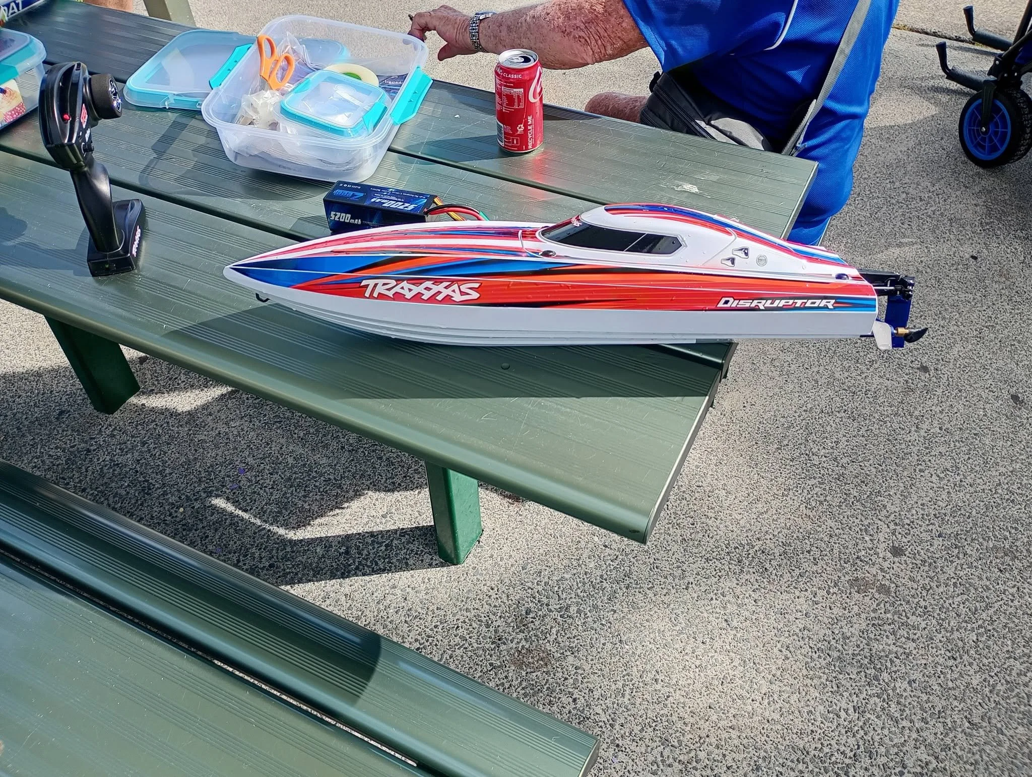 A remote-controlled boat labeled "Traxxas Disruptor" on an outdoor picnic table. There are containers, a soda can, a battery, and a remote control on the table. An elderly person in a blue shirt is sitting nearby, with a wheelchair visible in the bac