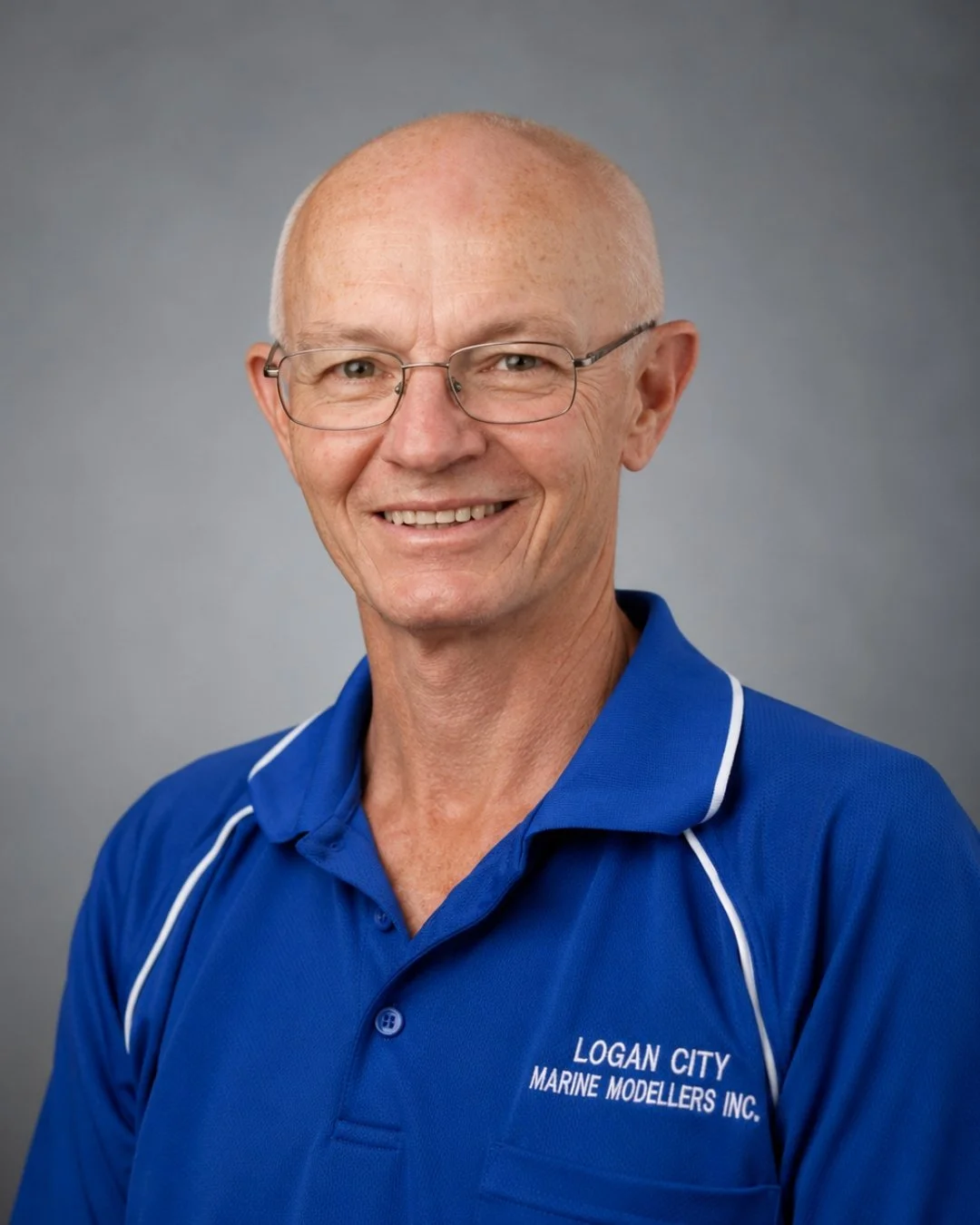 A smiling elderly man with glasses wearing a blue polo shirt with white piping that reads 'Logan City Marine Modellers Inc.'
